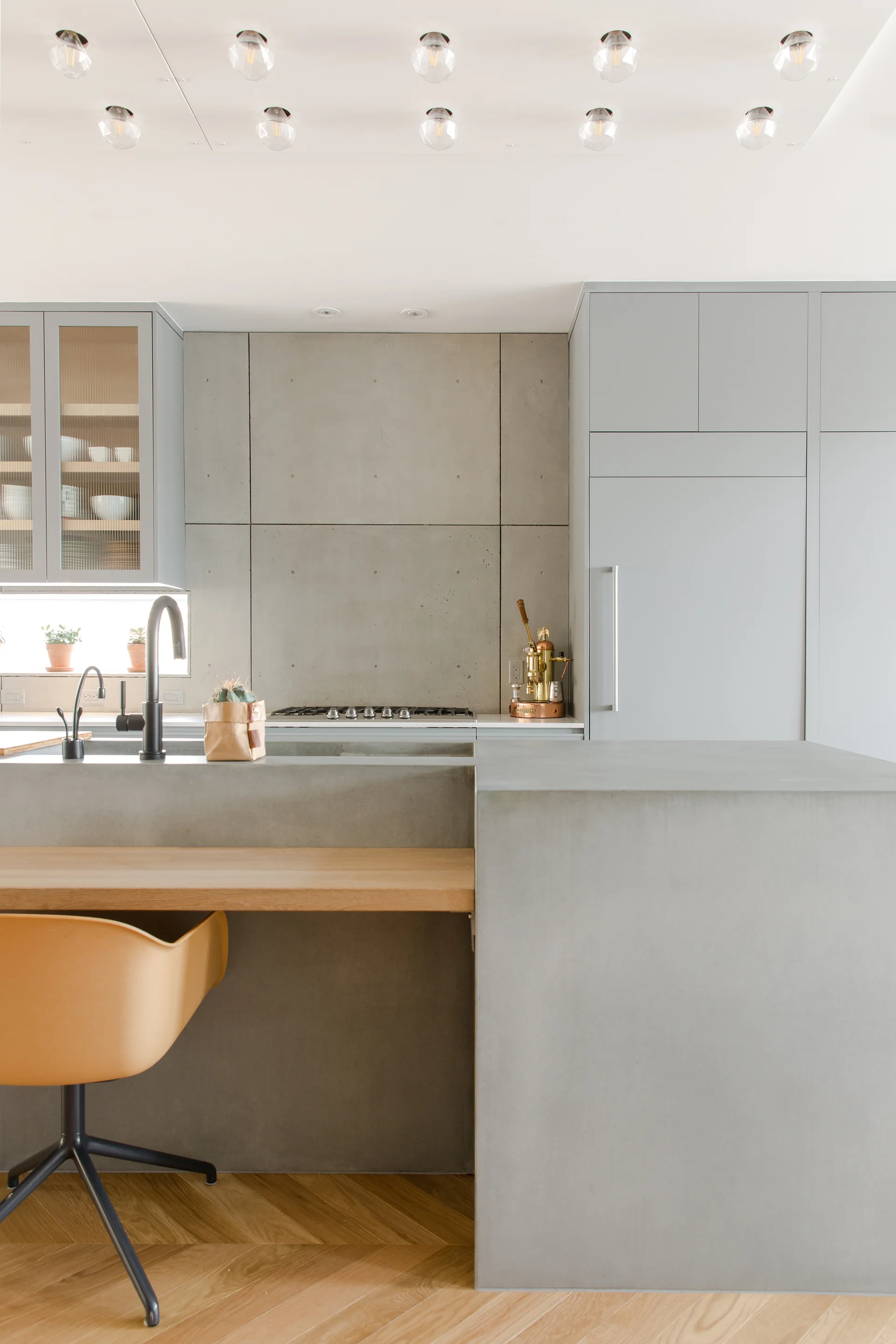 A modern kitchen featuring a gray concrete island with an integrated light wood breakfast bar, minimalist gray cabinets, and herringbone flooring.