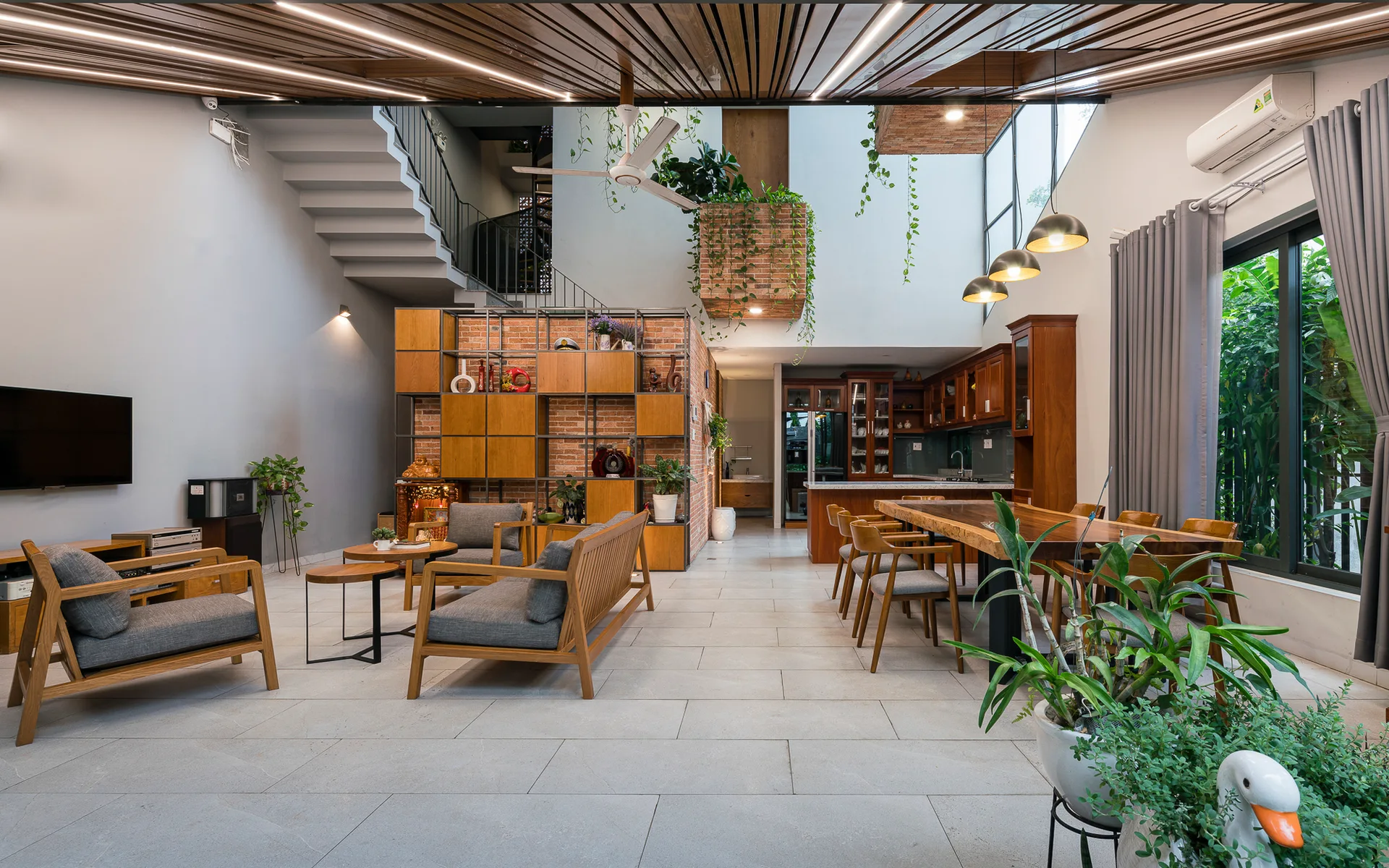 A modern open-plan living and dining area featuring a wood slat ceiling, concrete staircase, exposed brick wall partition, and wooden furniture.