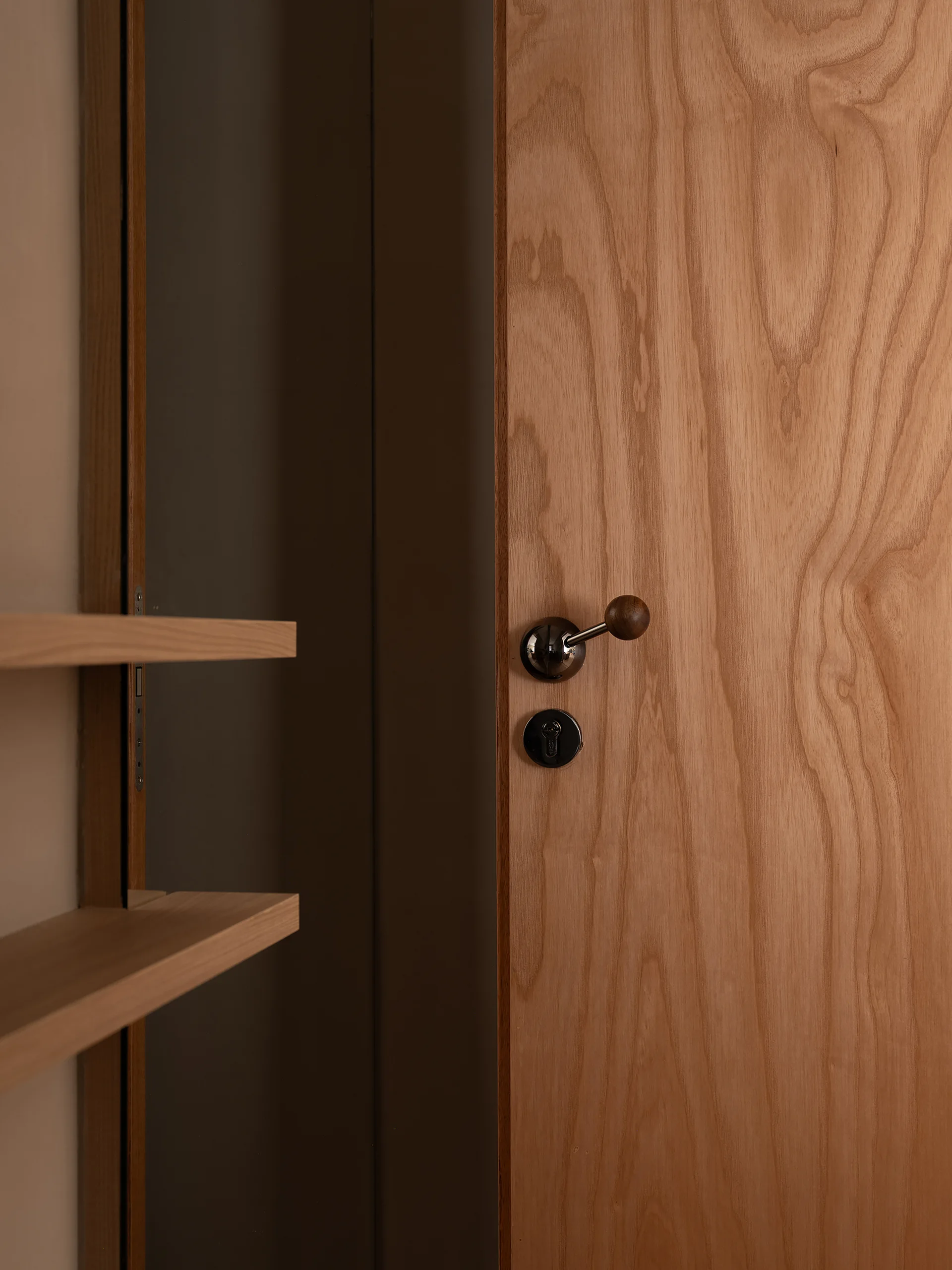 A close-up of a light wood door with an organic grain pattern featuring a metallic lever handle with a round wooden tip next to floating shelves.
