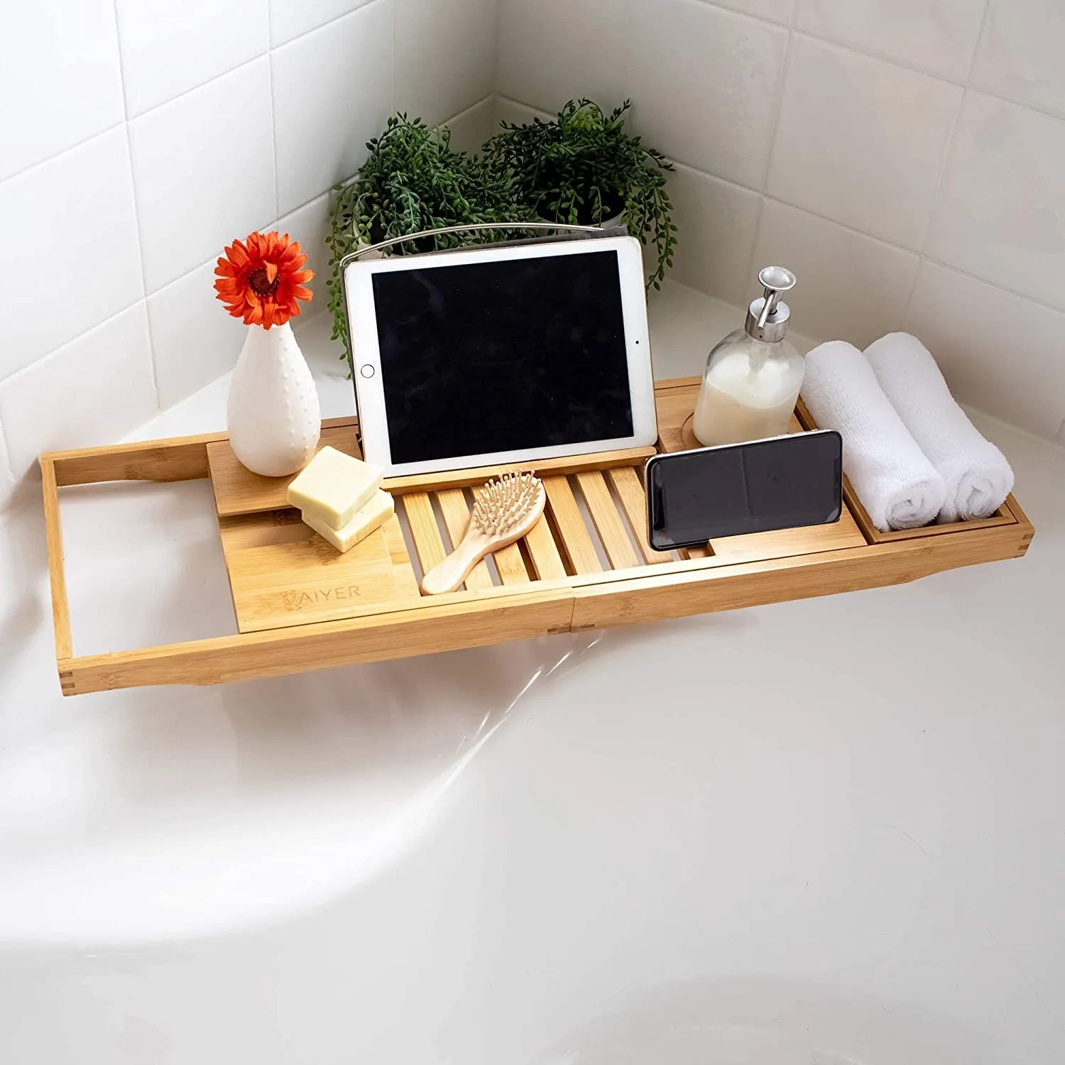 A slatted bamboo bathtub tray spans a white tub, holding a tablet, smartphone, rolled towels, and a small flower vase against white tile.