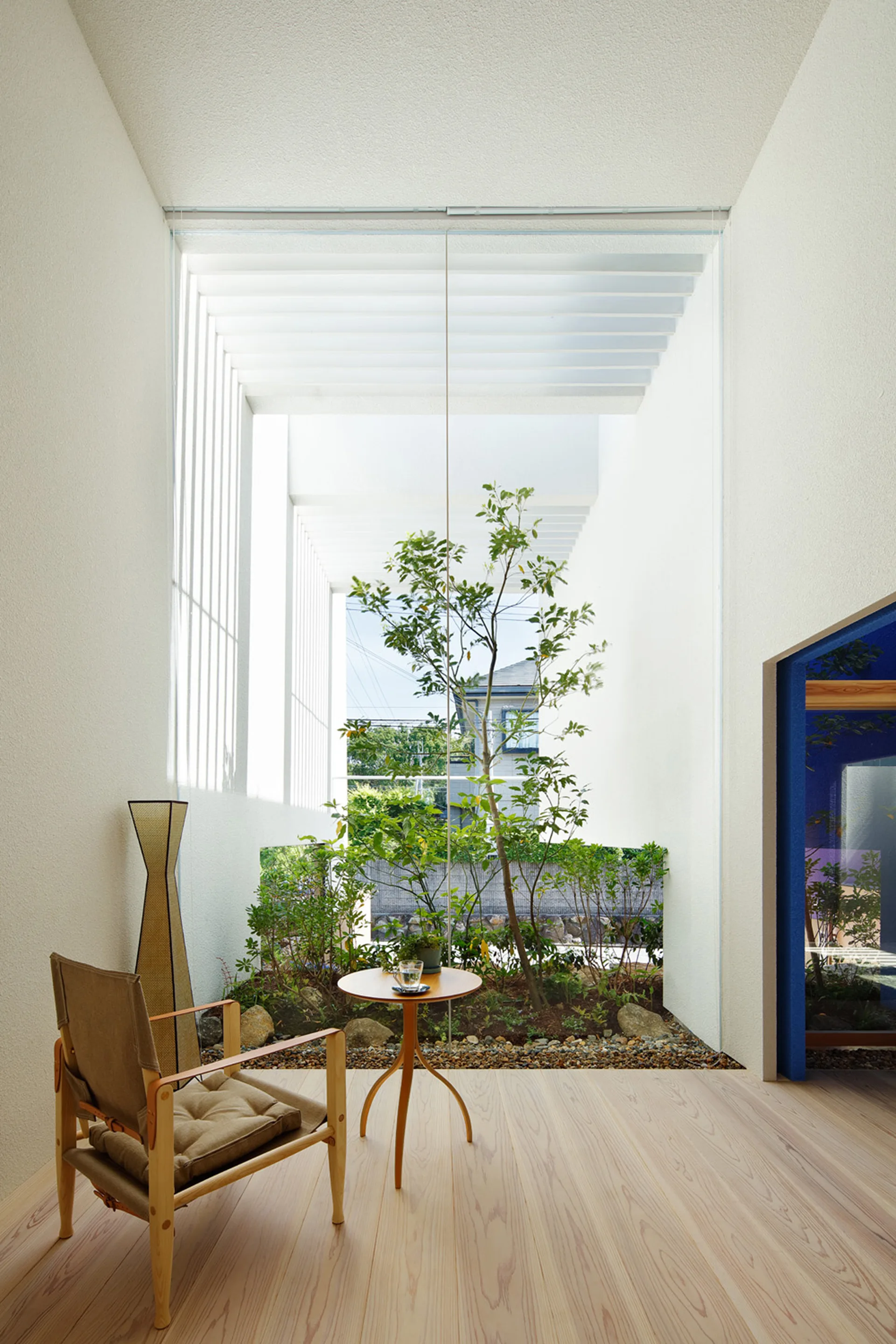 A minimalist sitting room features a wood armchair and round table next to a large glass wall displaying an outdoor pocket garden.