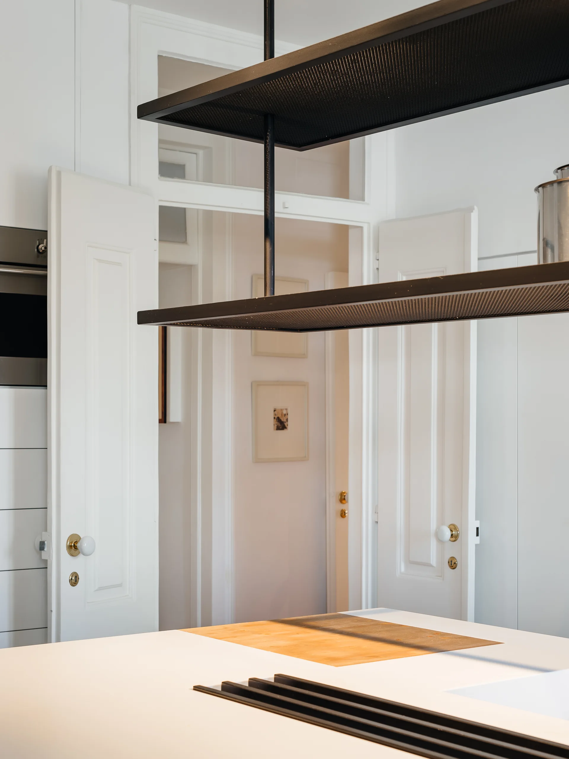 A white kitchen island features an inlaid wood surface beneath a suspended black metal rack, with white double doors and brass hardware in the background.