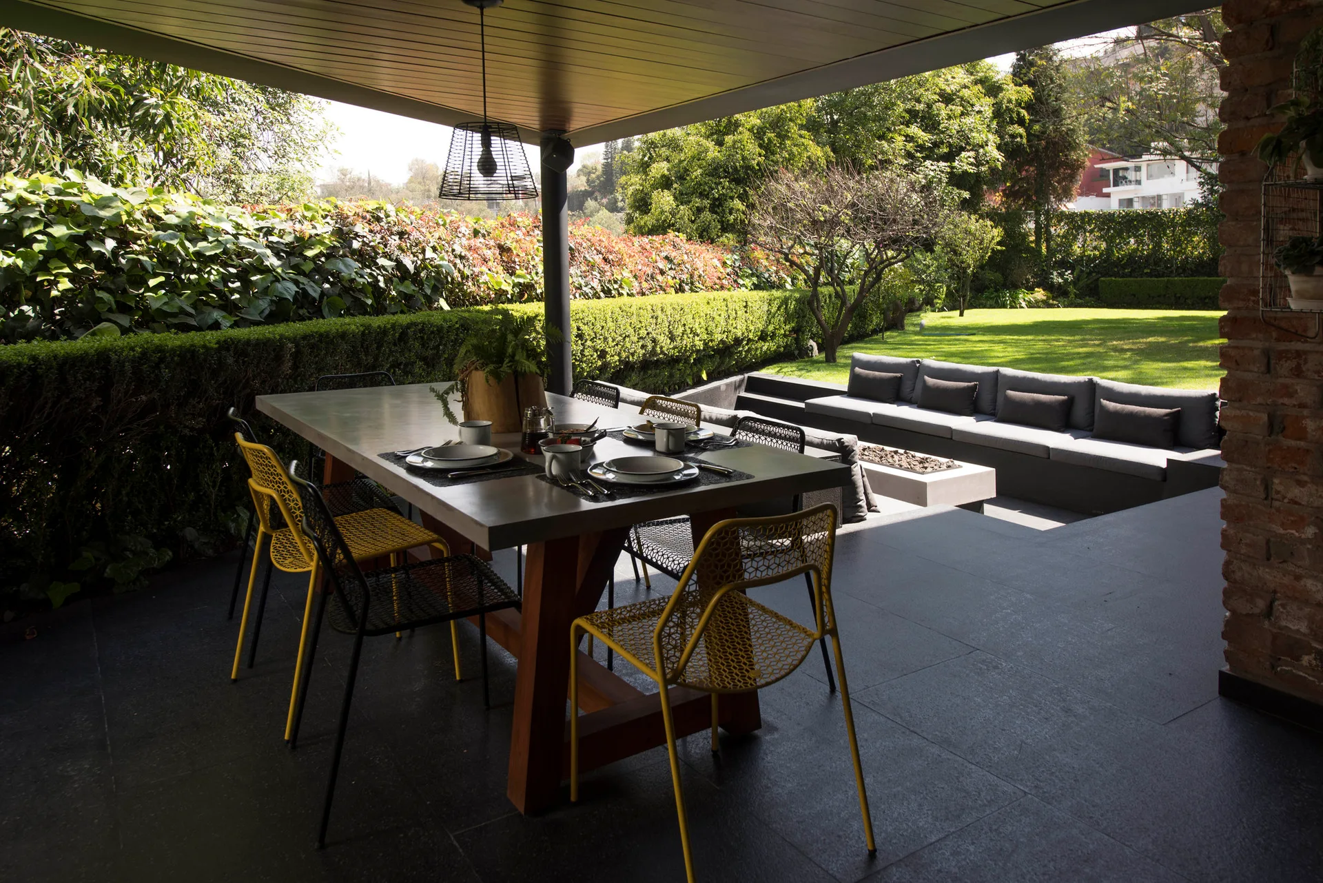 A covered patio dining table with yellow and black chairs sits next to a sunken outdoor lounge with a gray sectional and garden views.