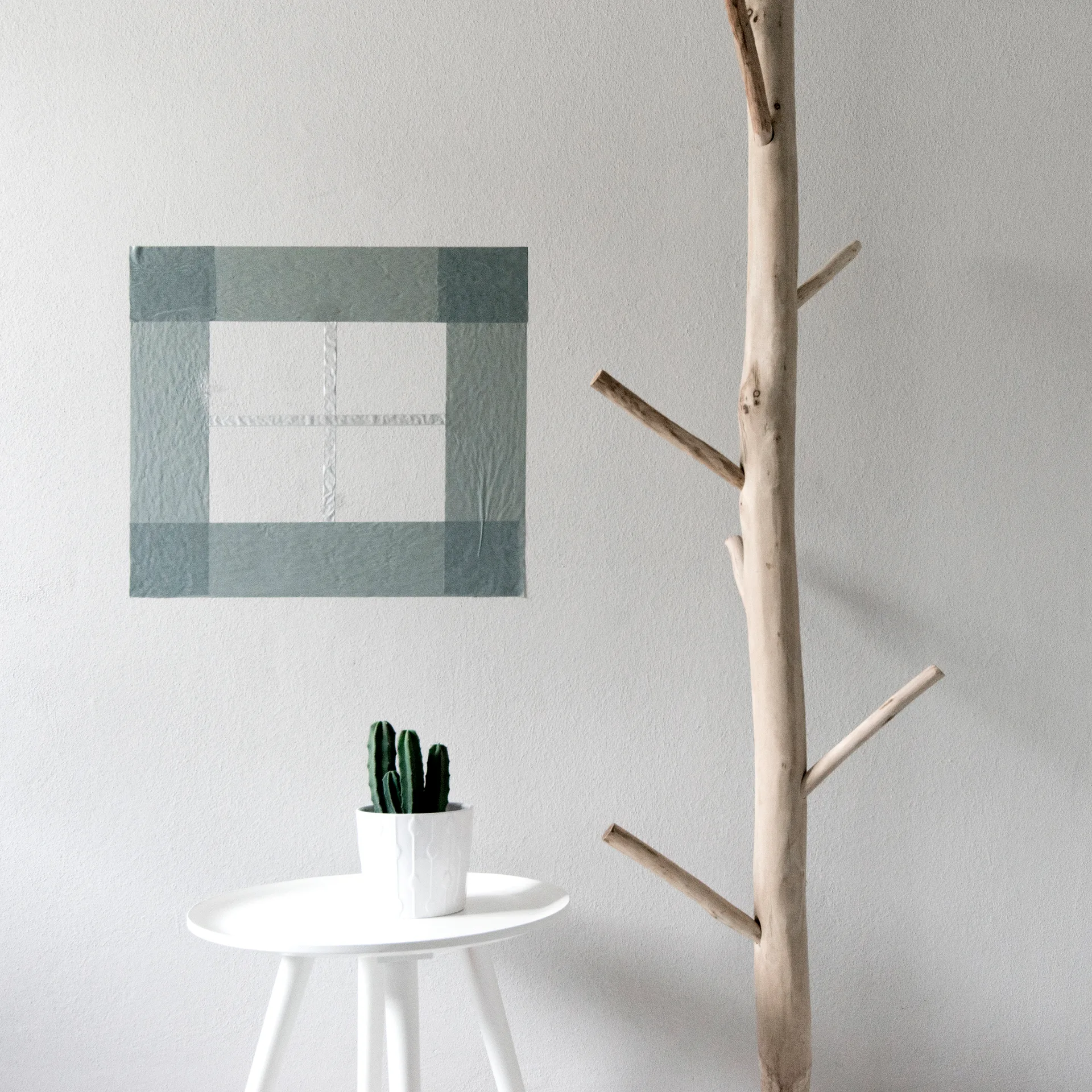 A natural wood branch coat rack stands next to a white pedestal table holding a potted cactus against a grey wall with a square paper collage.