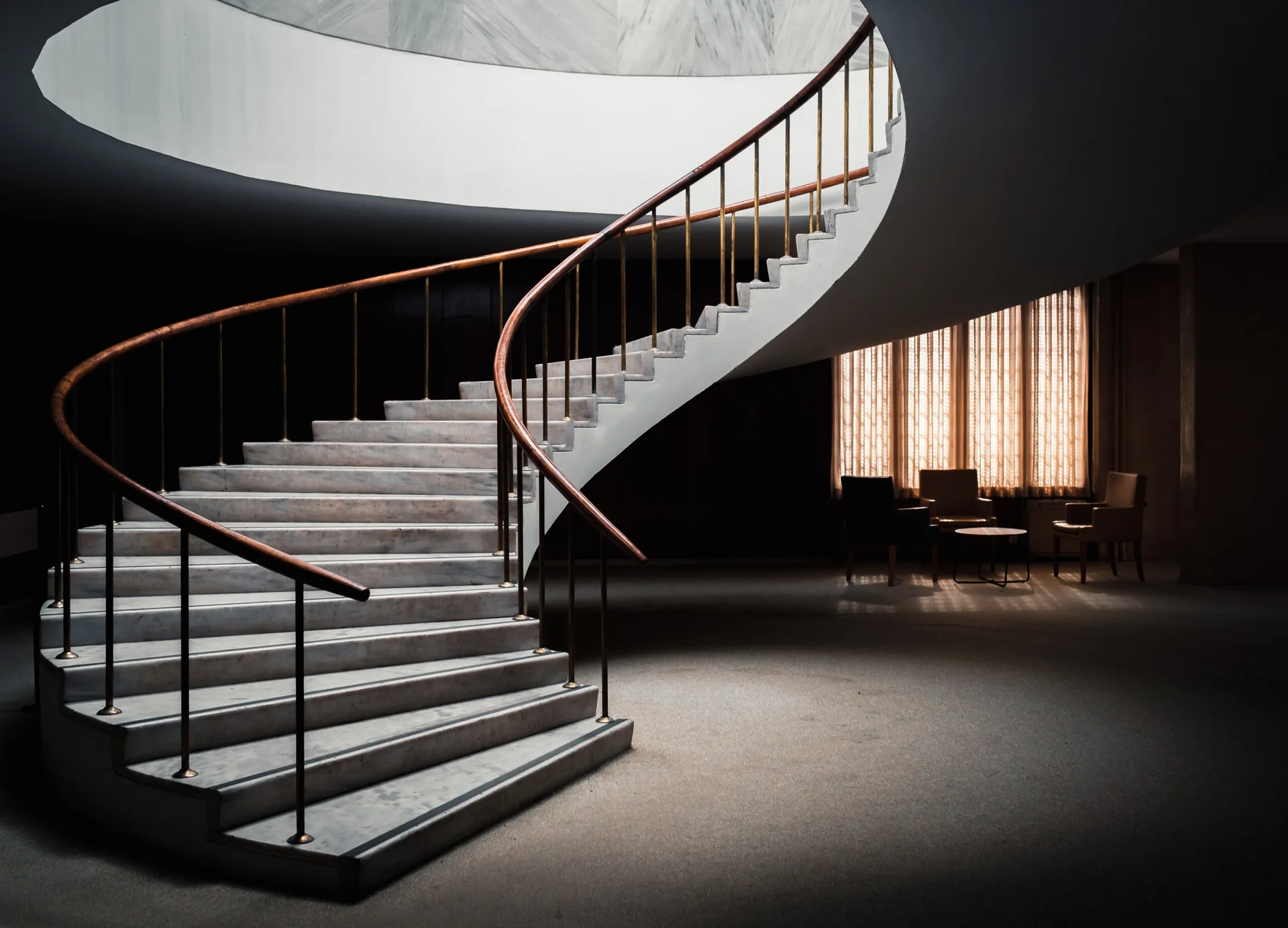 A grand marble spiral staircase with a curved wooden handrail and brass spindles stands in a dimly lit hall with armchairs near a window.