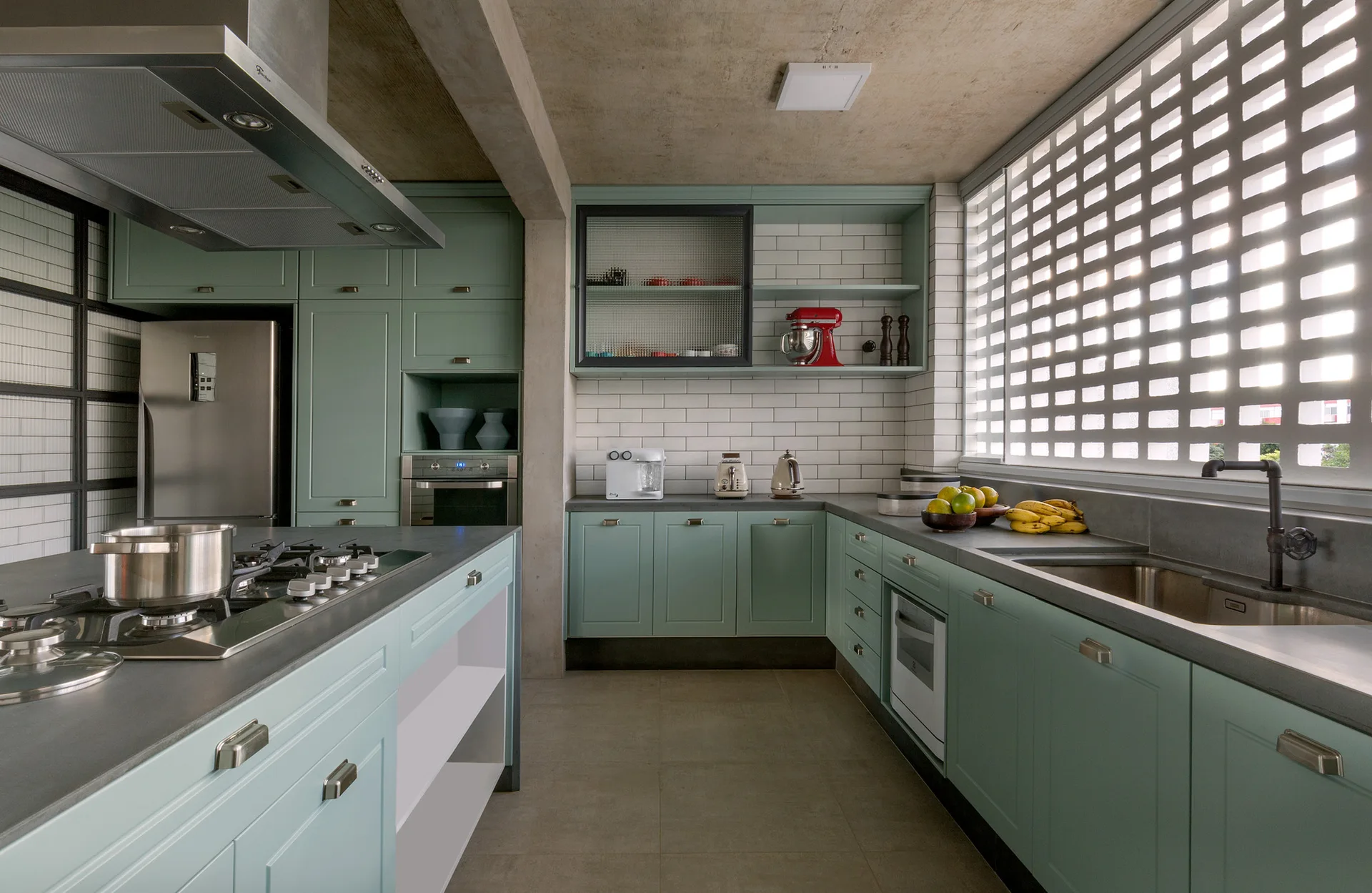 A modern kitchen featuring sage green cabinetry, a large window with a white grid screen, concrete ceiling, and a central island with a cooktop.