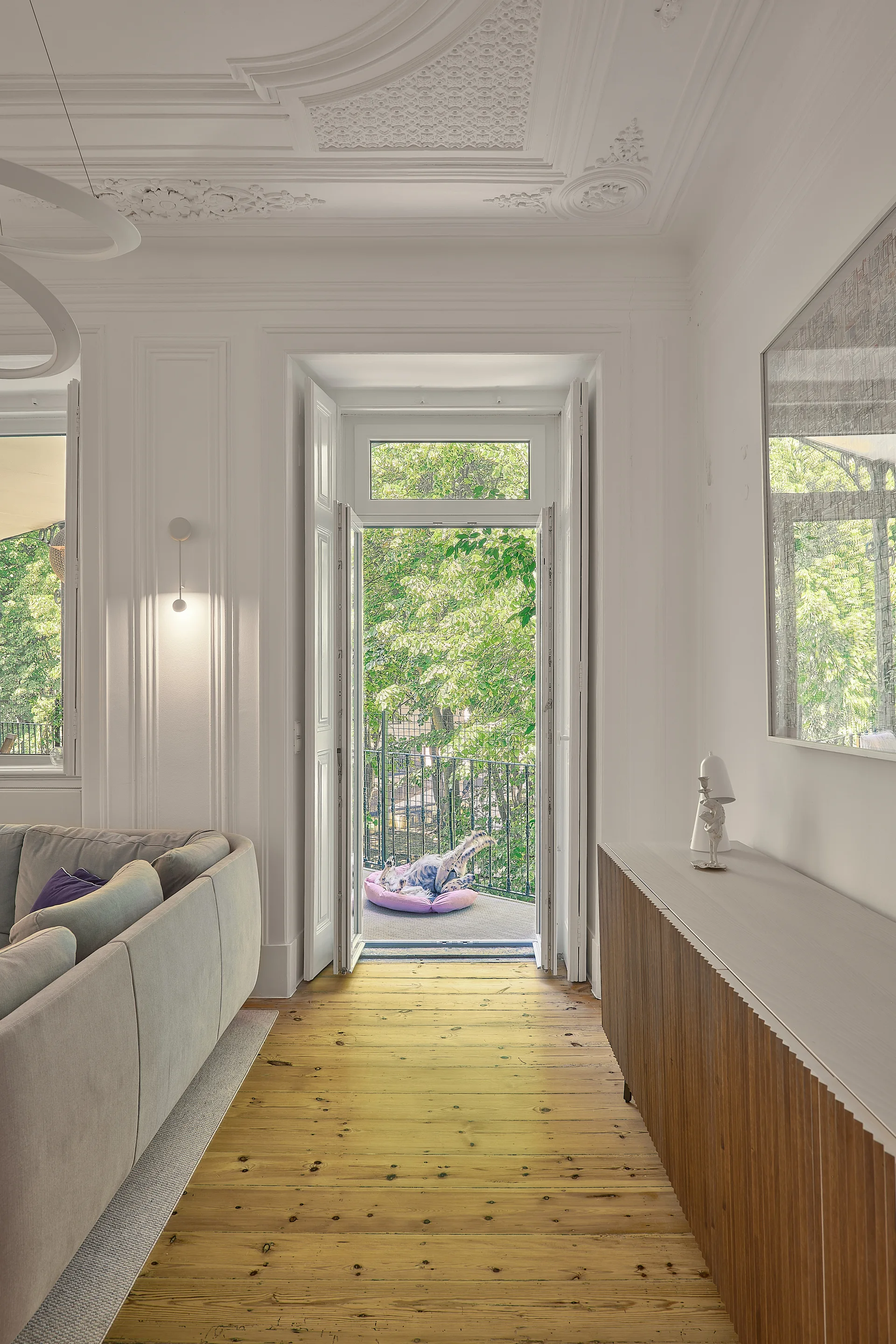 A bright living room features a grey sectional sofa, a fluted wood sideboard, and an ornate plaster ceiling leading to open balcony doors.