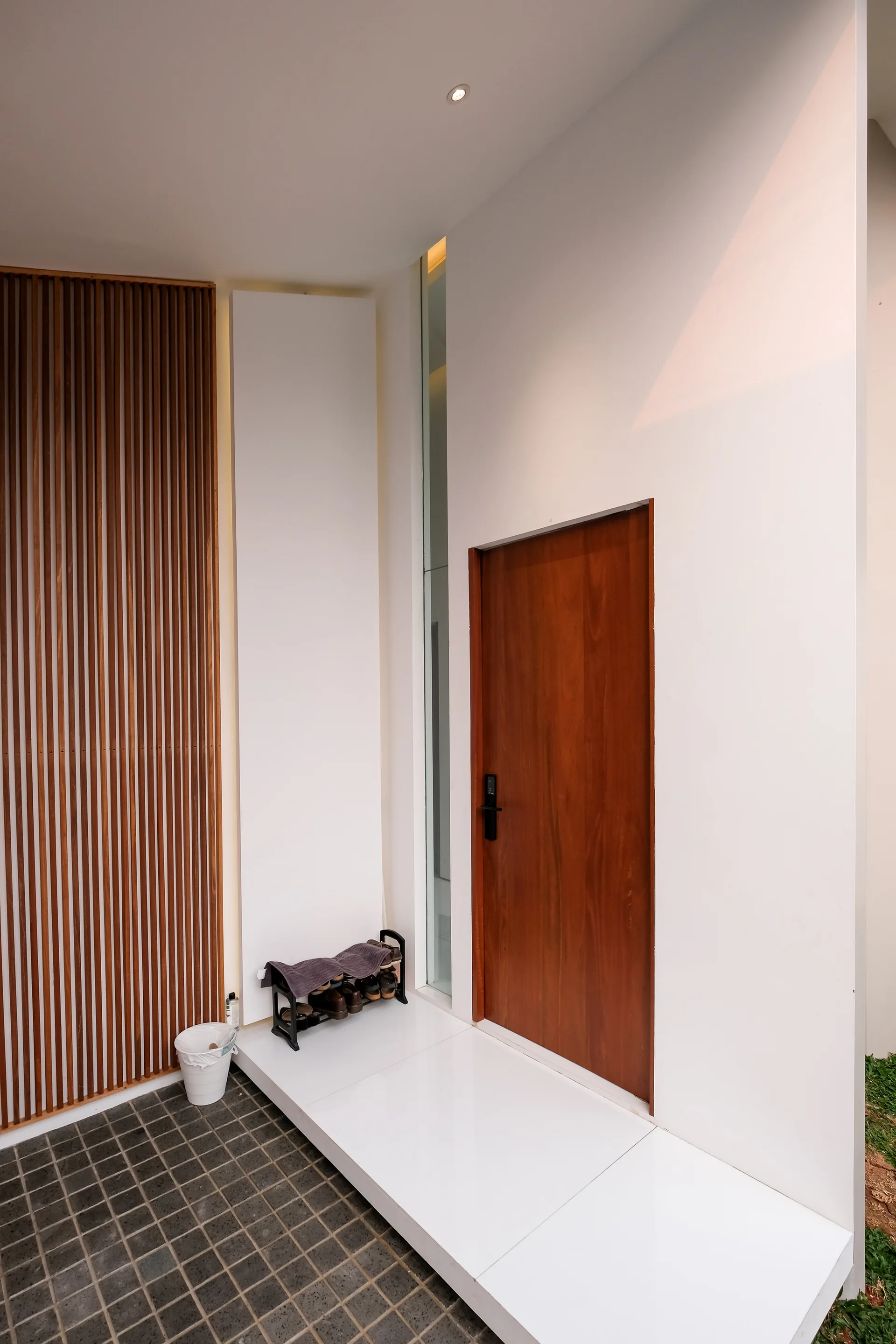 A modern entryway features a rich wood door, a narrow vertical glass window, white walls, and a decorative wood slat panel beside a raised tiled floor.