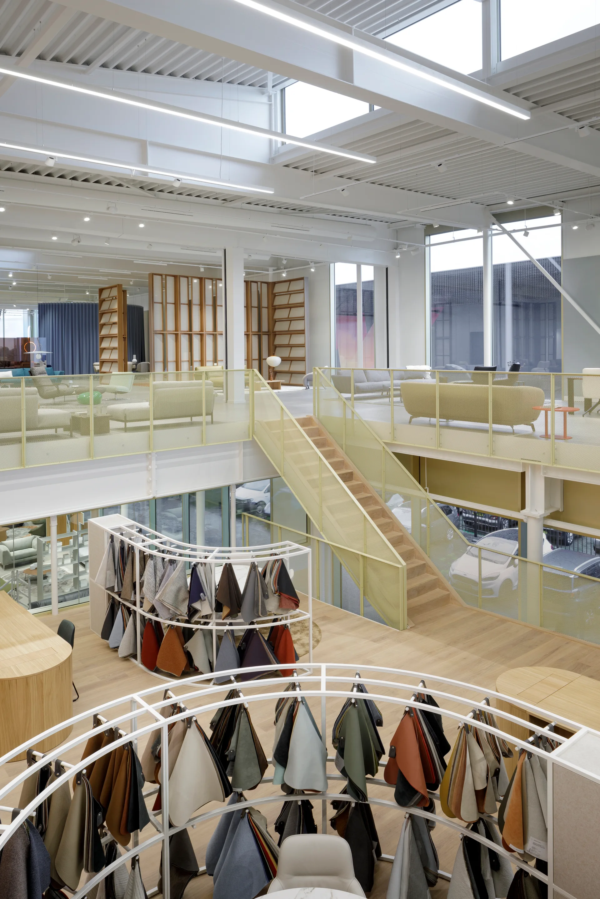A multi-level design showroom featuring curved metal racks with hanging fabric samples on the wood floor and a staircase with a yellow mesh railing.