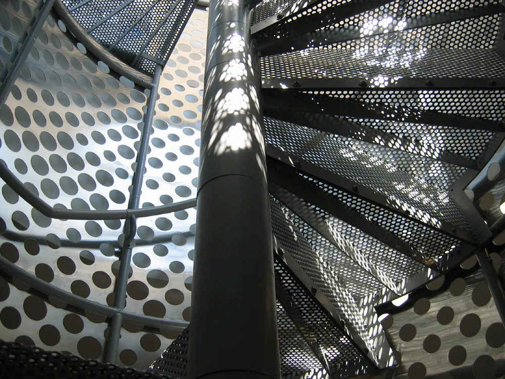 A close-up view of a metallic spiral staircase featuring perforated metal steps wrapping around a central support column.