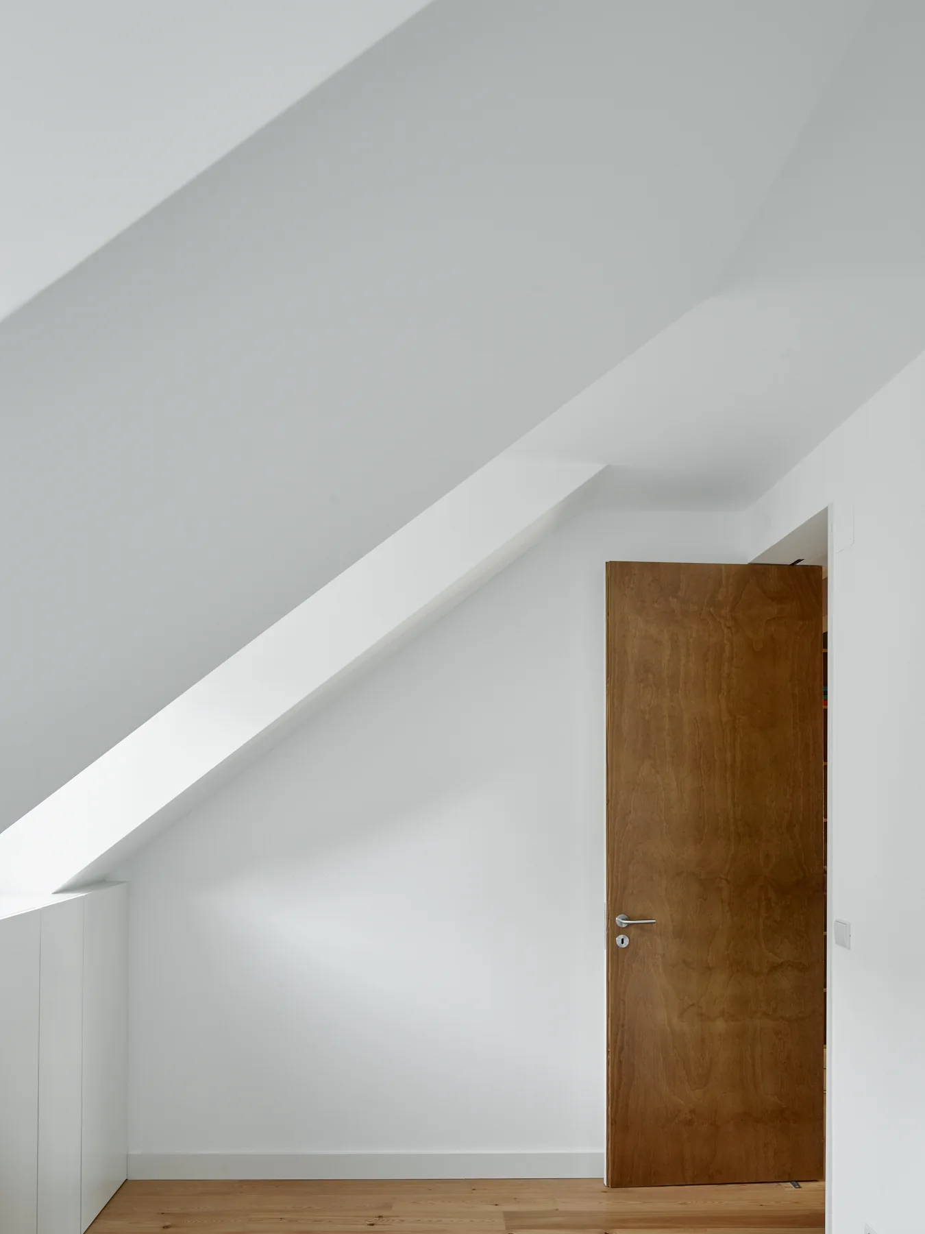 A minimalist attic room features smooth white walls, a steeply pitched ceiling, light wood floors, and a simple warm wood door.
