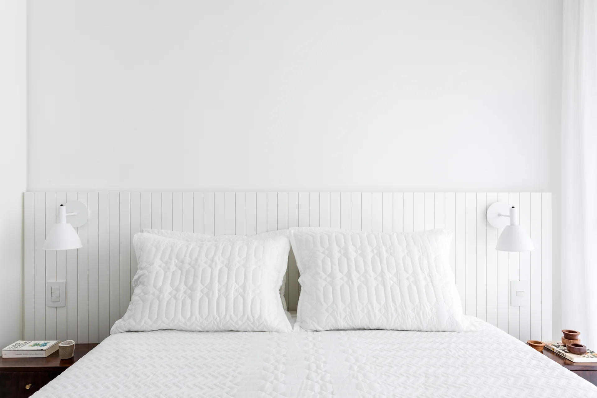 A symmetrical minimalist white bedroom featuring a bed with quilted pillows set against a vertical slatted headboard wall with matching sconces.