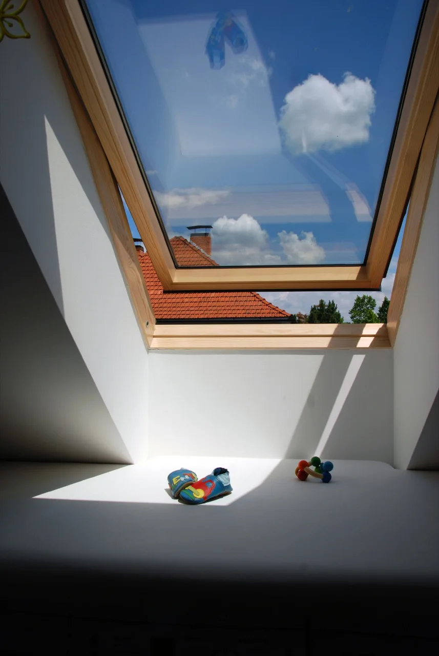 An open wooden-framed skylight in a white attic wall reveals a blue sky, while colorful baby shoes and a toy rest on the sunny windowsill below.