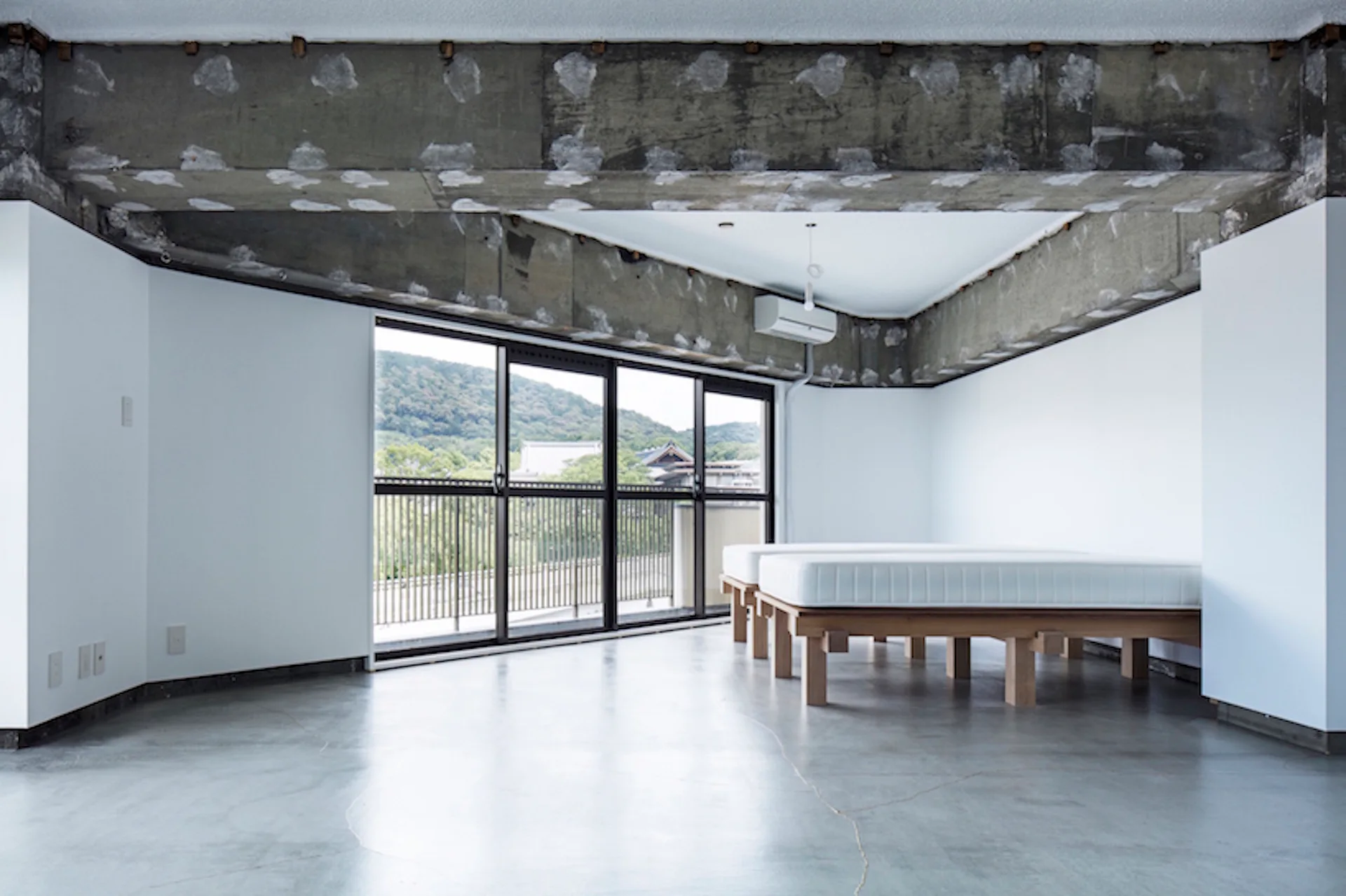 A minimalist bedroom featuring two low wooden platform beds on a polished concrete floor beneath a rough exposed concrete ceiling beam.