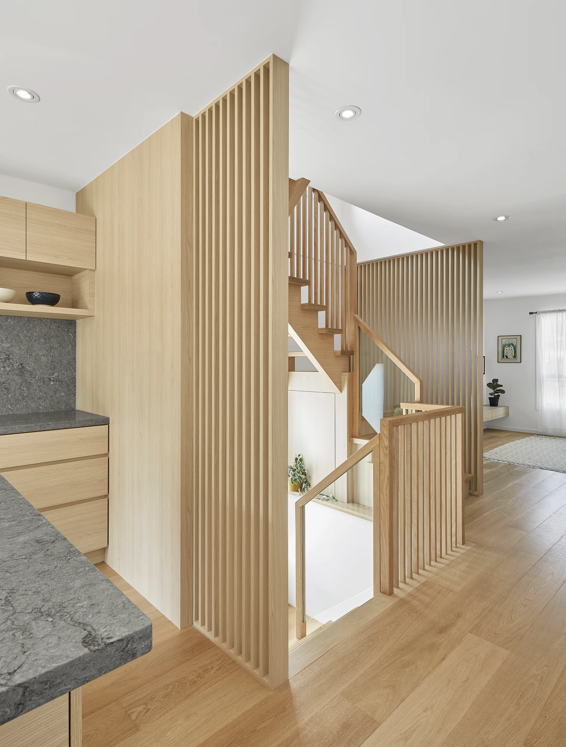 A modern light wood staircase features vertical wooden slat screens and a glass railing next to kitchen cabinets and grey countertops.