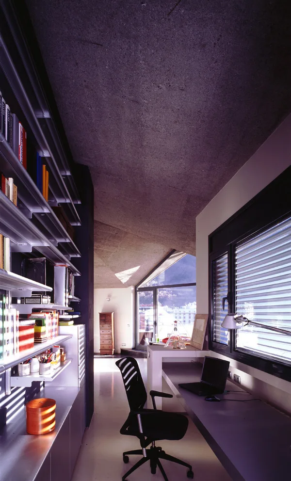 A modern home office features a long built-in desk, a black ergonomic chair, and metal shelving beneath a sloped raw concrete ceiling.