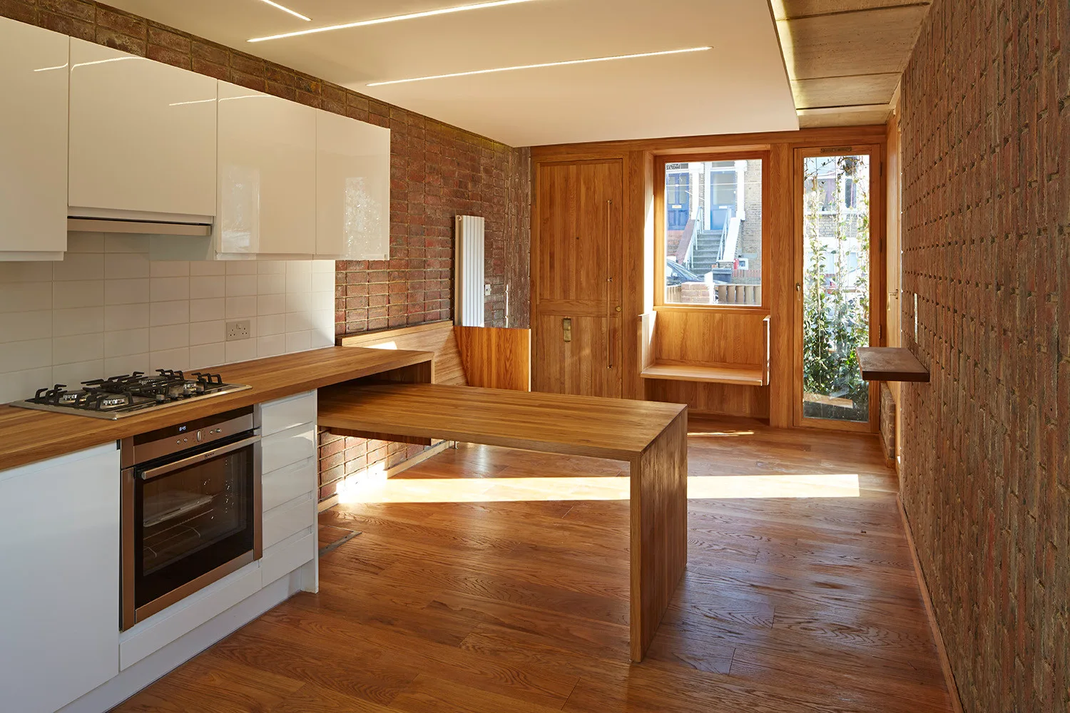 A modern kitchen with high-gloss white cabinets, a wooden dining table extension, and exposed brick walls.