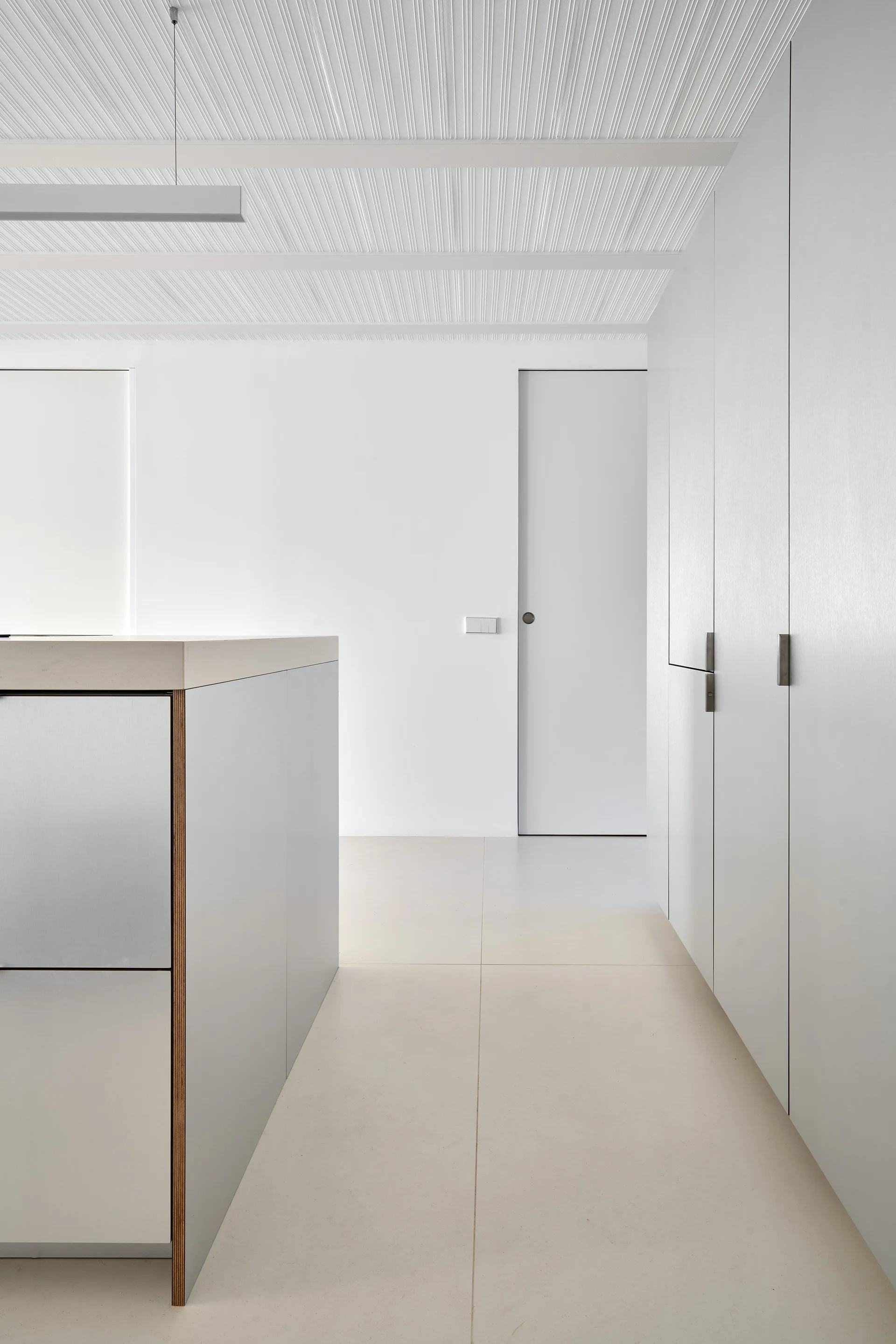 A minimalist kitchen features a metallic front island with a thick countertop, seamless white tall cabinets, and a grooved, textured ceiling.