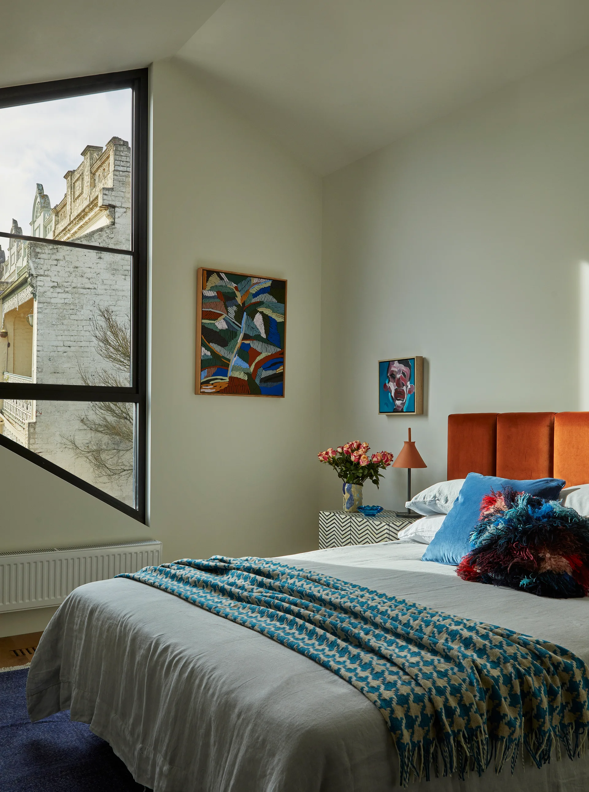 A bedroom featuring an angled black-framed window, a rust-orange upholstered headboard, and a chevron-patterned nightstand next to the bed.