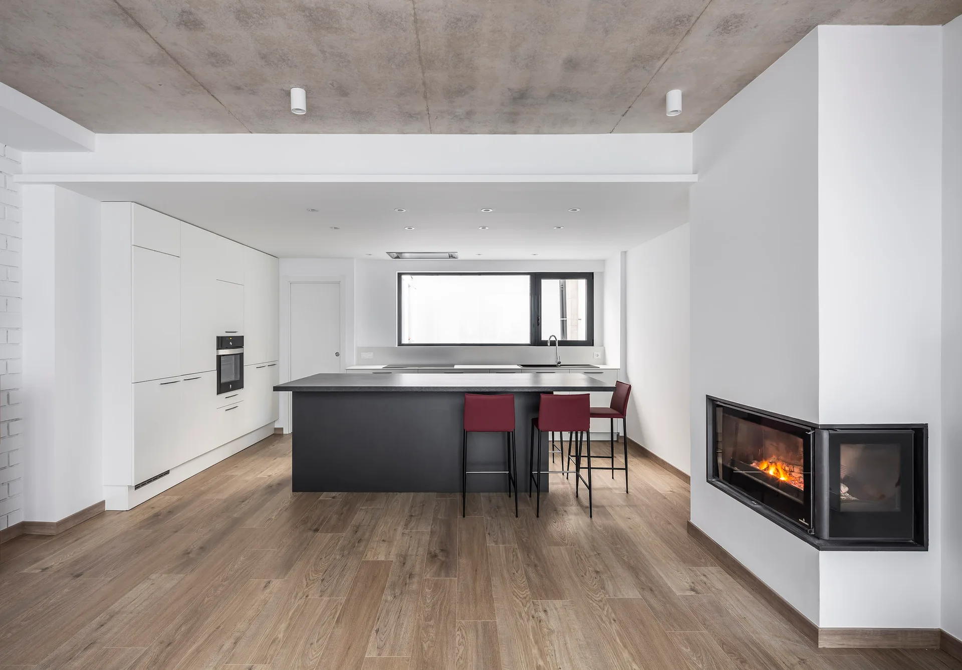 A modern kitchen with white flat-panel cabinets, a large black central island with three red stools, and an exposed raw concrete ceiling.