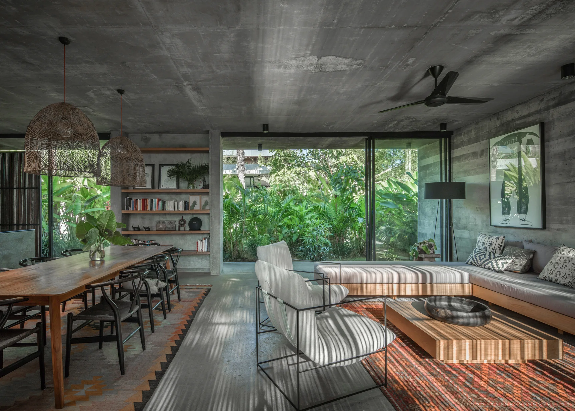 An open living and dining area with exposed concrete ceilings, a long wood dining table, and a modular sofa looking out at a lush garden.