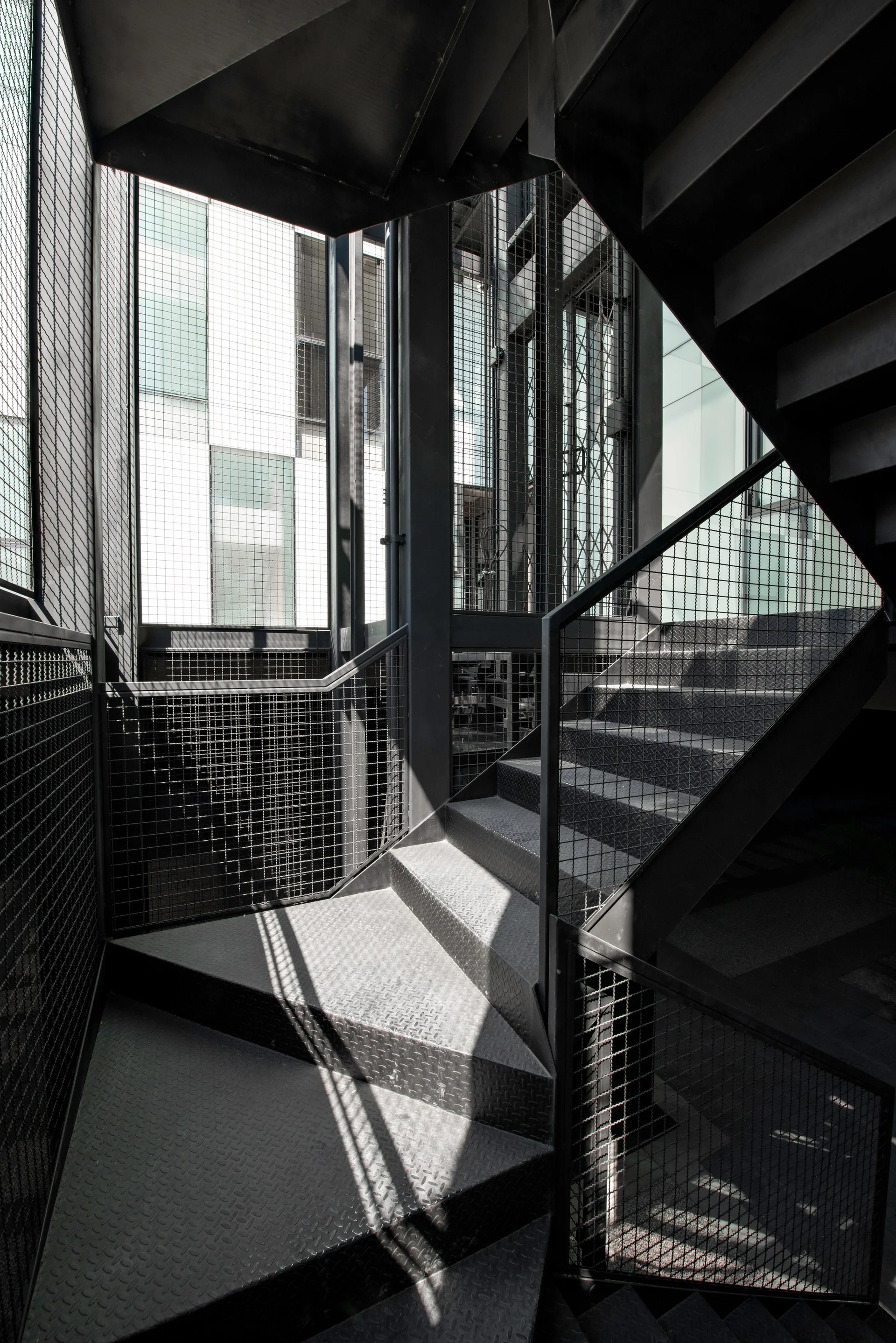 A black metal industrial staircase features checkered diamond plate treads and wire mesh safety railings illuminated by natural sunlight from a window.