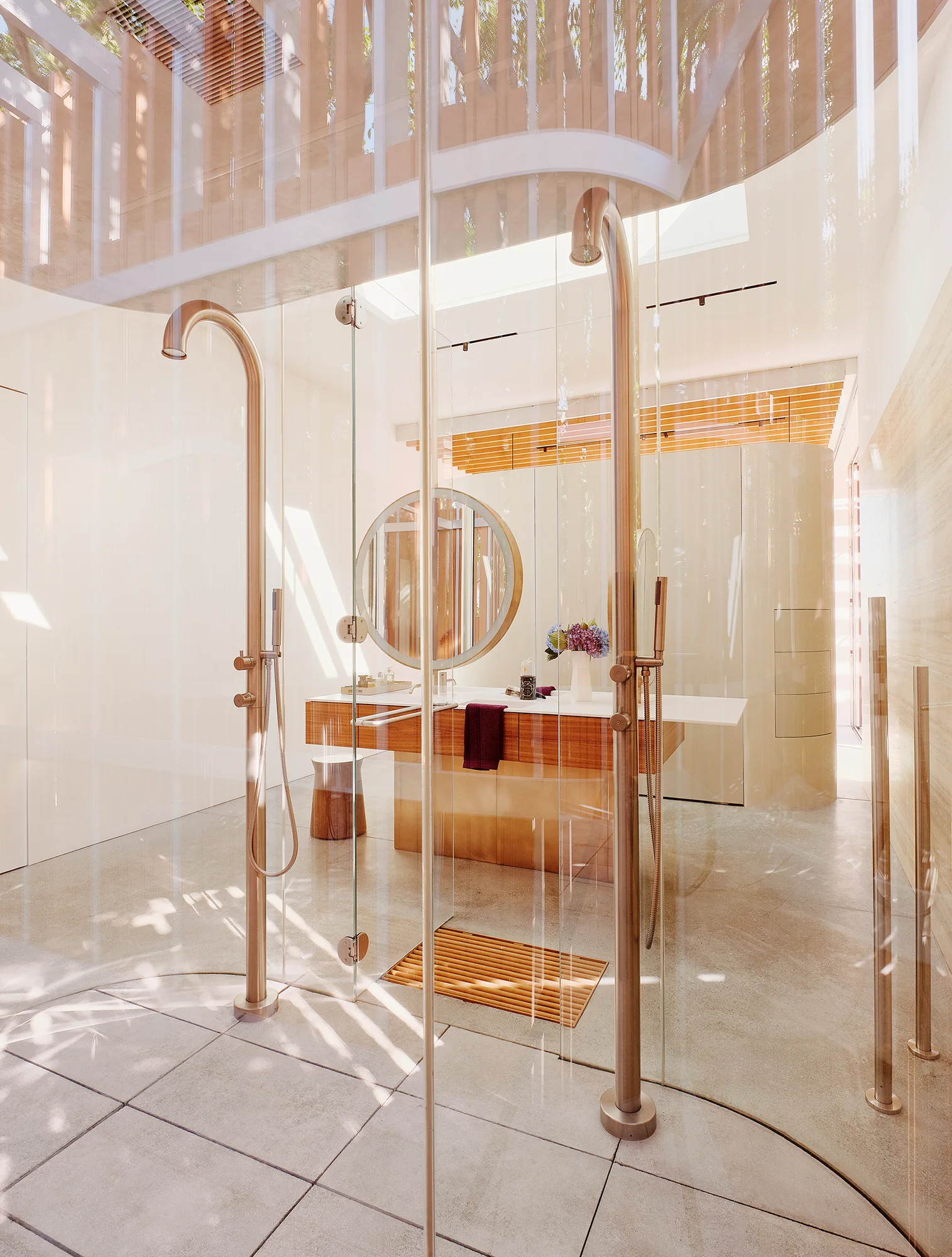 A modern beige bathroom features dual freestanding metal shower heads behind glass partitions and a central wood vanity with a round mirror.