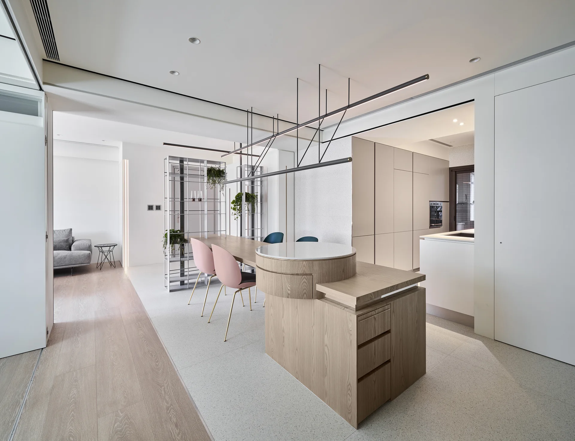 A minimalist kitchen features a light wood island with an integrated dining table, pink chairs, and a sleek black linear pendant light.