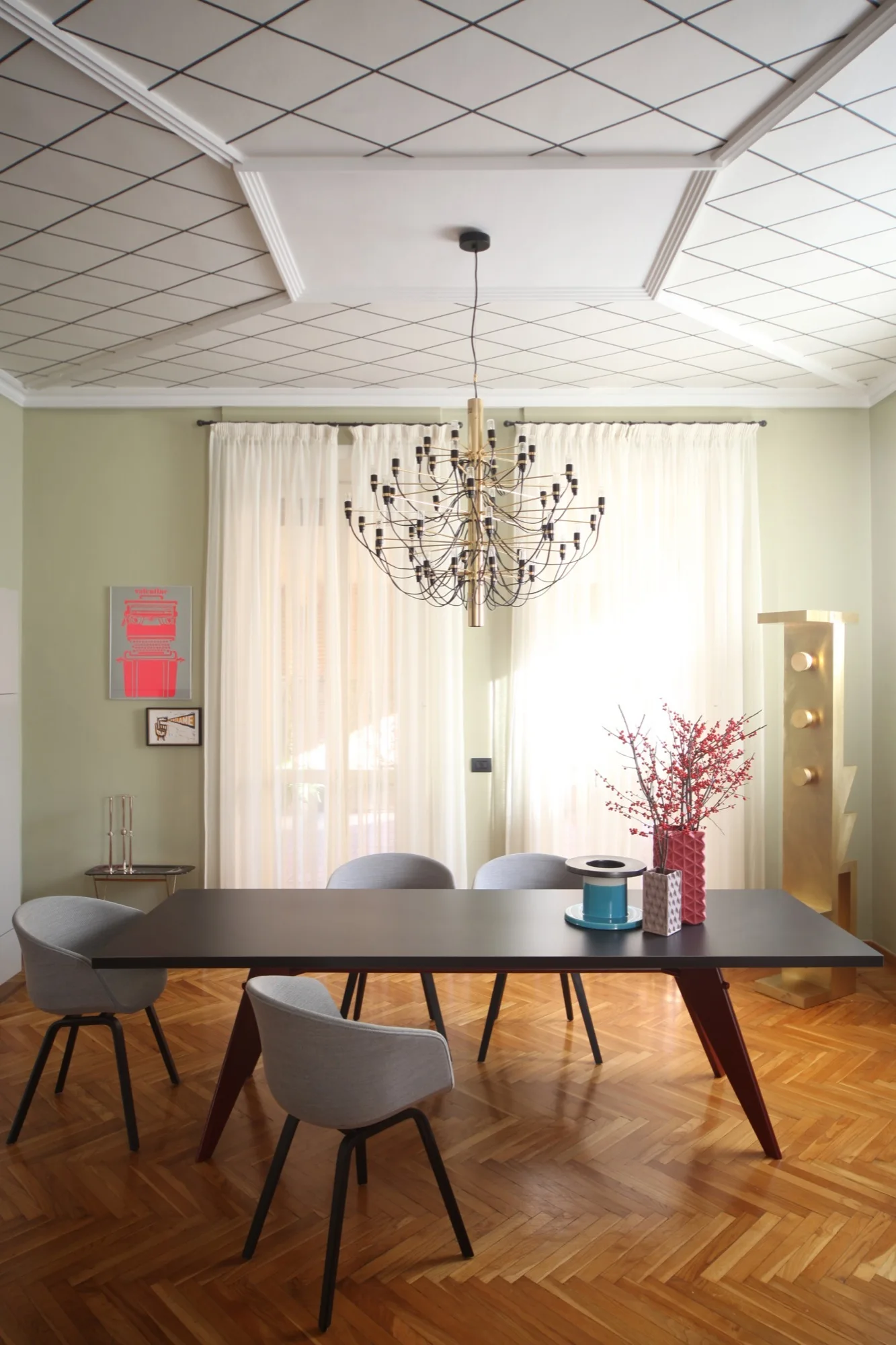 A modern dining room featuring a dark table, grey chairs, a branching metal chandelier, and a white ceiling with a black diamond grid pattern.