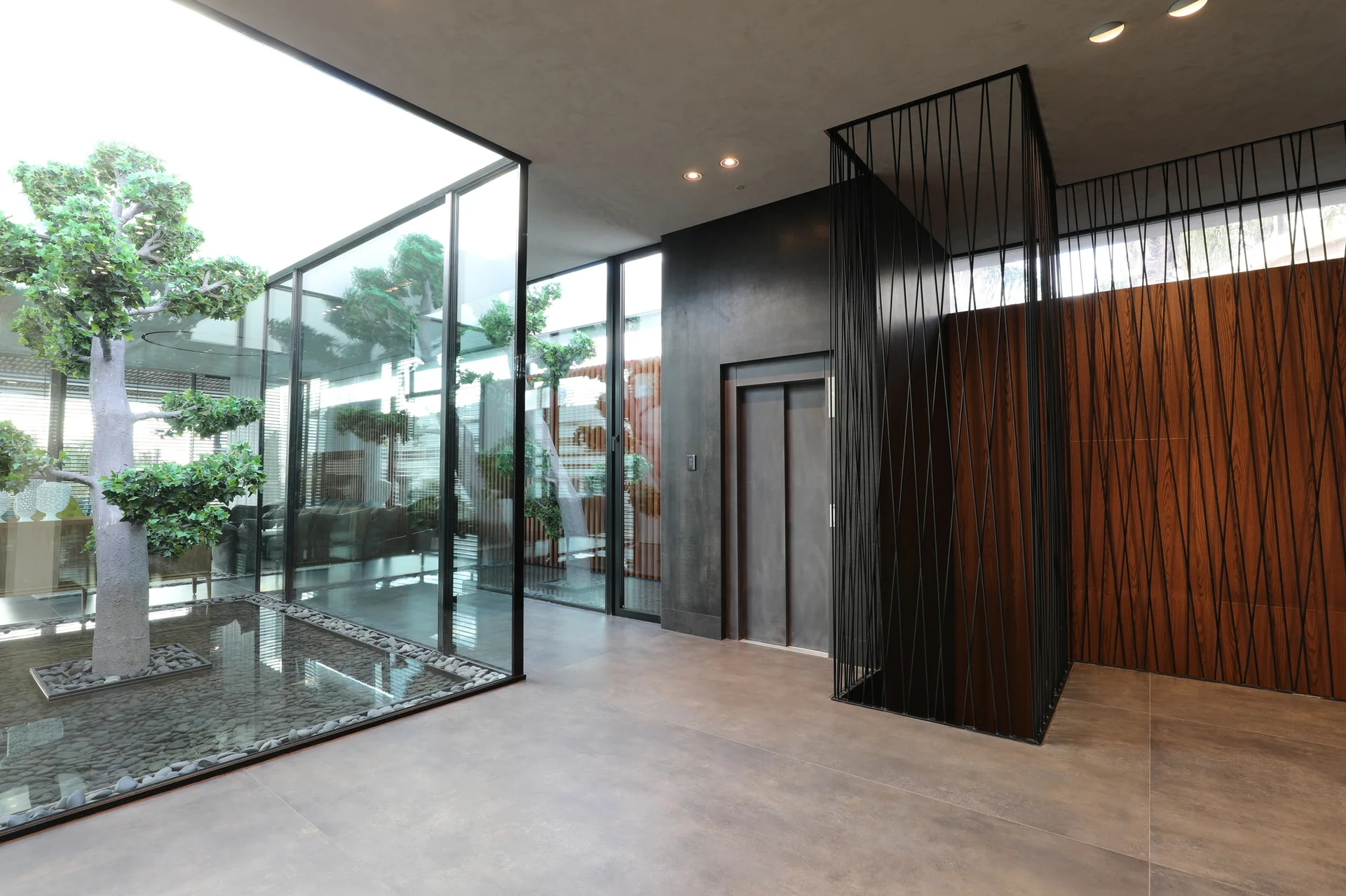 A modern entryway features a glass-walled indoor courtyard with a tree alongside a metallic elevator framed by vertical black metal rods.