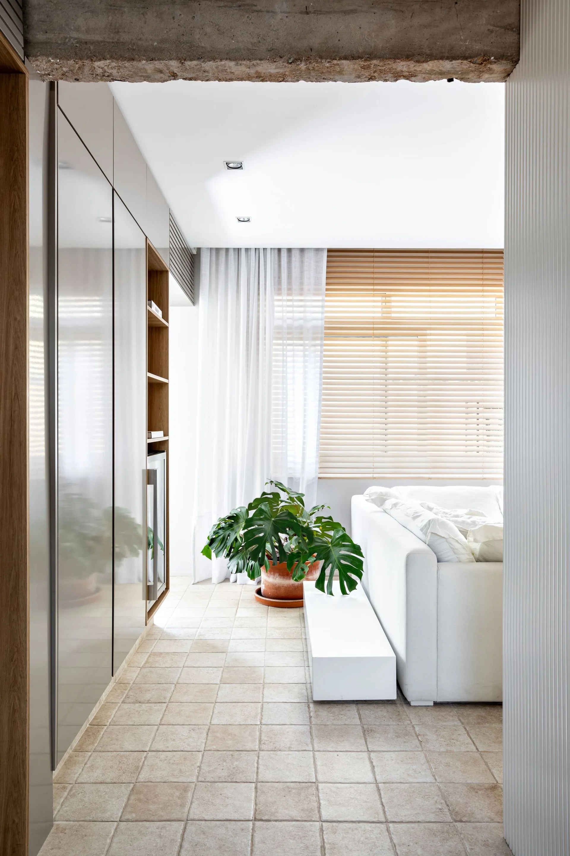 A bright living space featuring a white sofa, neutral tiled floors, sleek built-in cabinets, and a window with wooden blinds and sheer curtains.