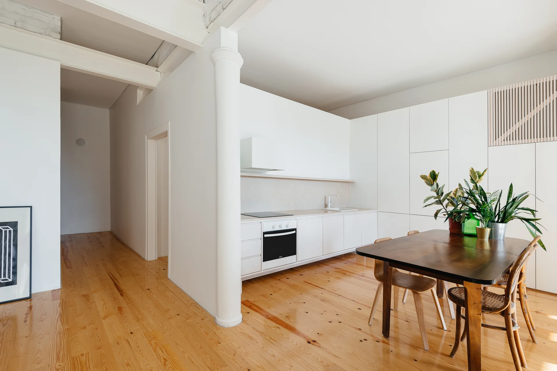 A minimalist white kitchen and dining area featuring a structural column, warm wood floors, and a dark wood table.