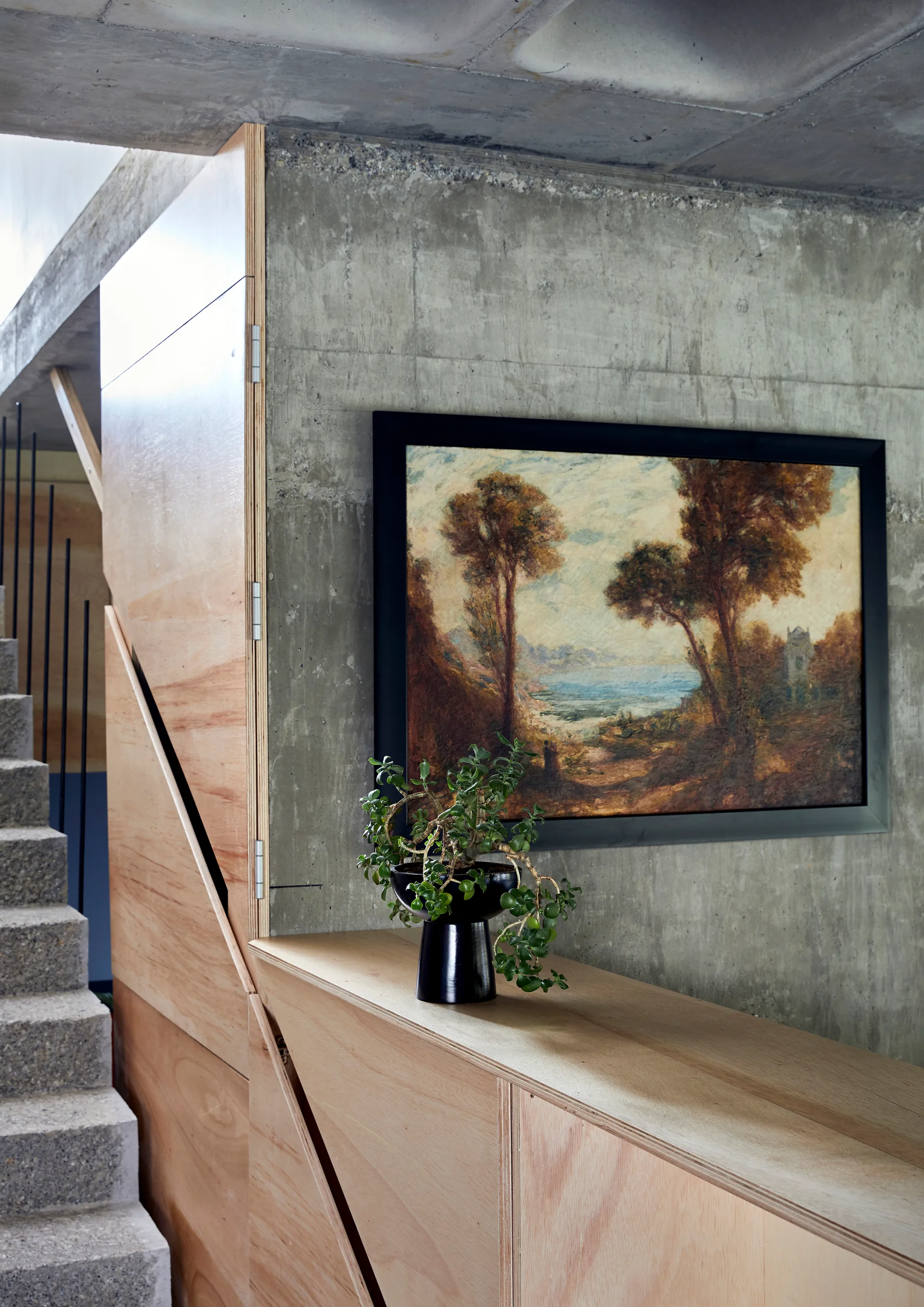 A concrete staircase stands next to built-in light wood cabinetry under a raw concrete wall displaying a framed landscape painting and a potted plant.
