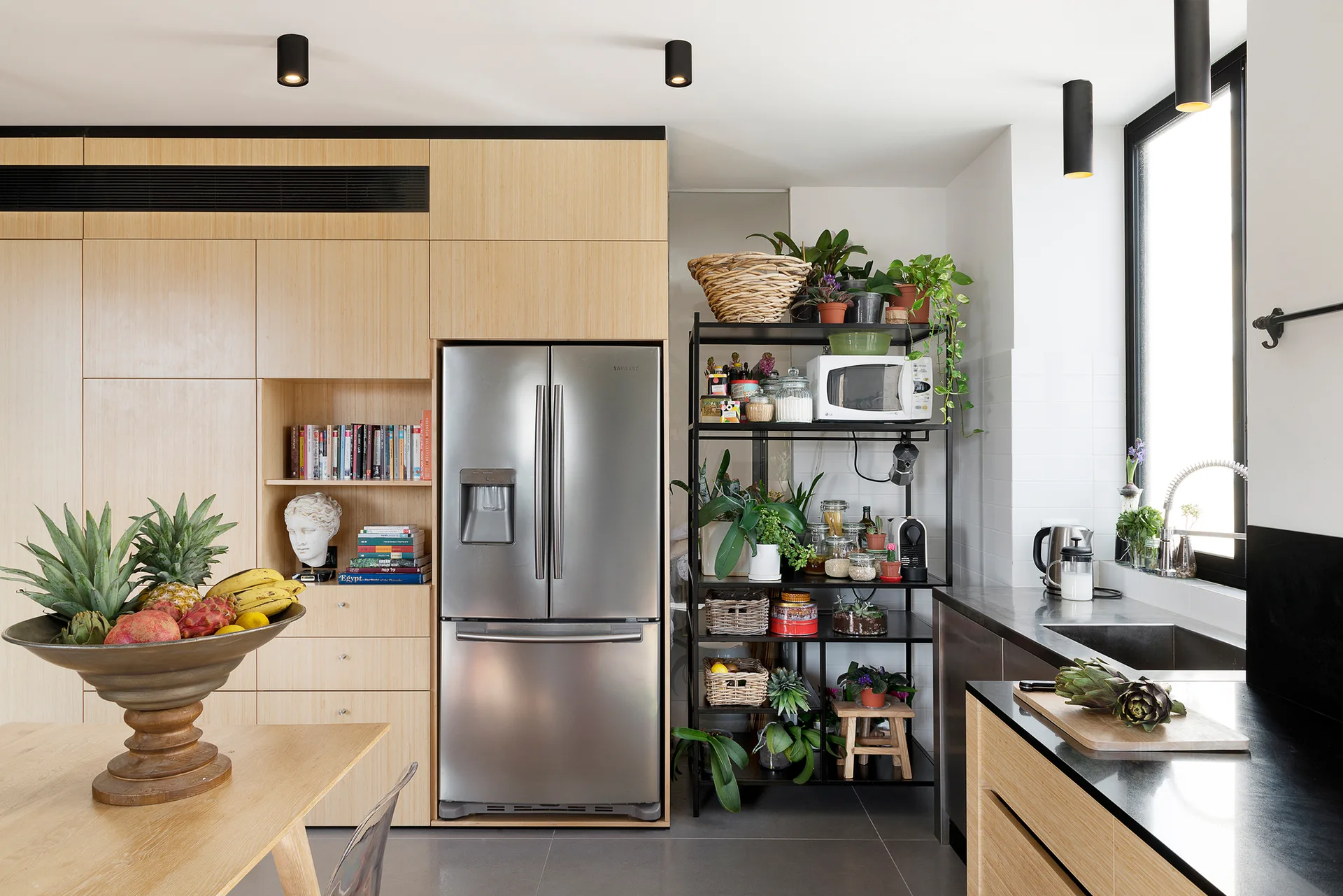 A modern kitchen with a light wood cabinet wall, stainless steel refrigerator, black metal shelving unit holding plants, and dark countertops.
