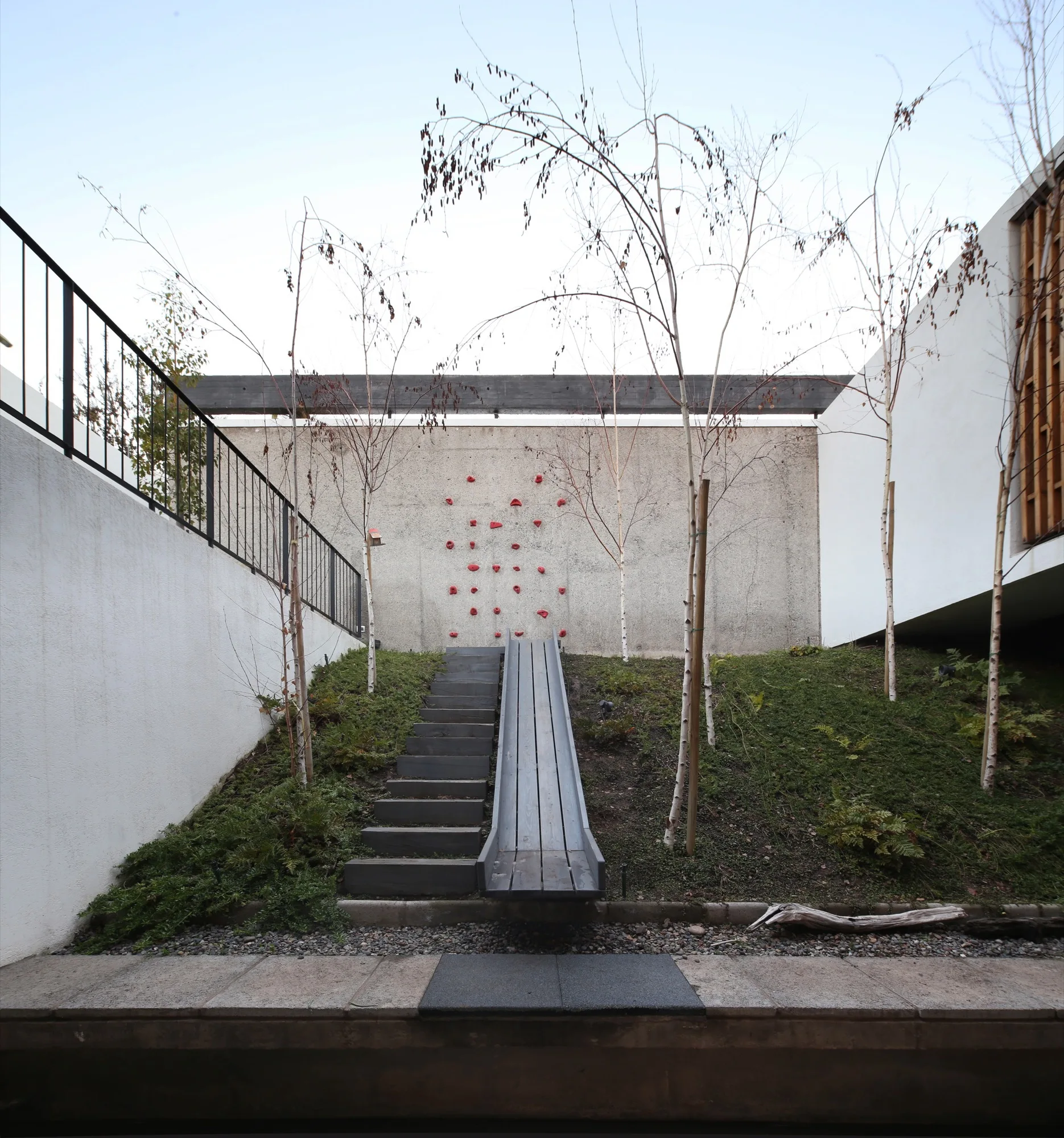 A terraced courtyard garden features a dark wooden slide and stairs alongside a concrete wall equipped with red climbing holds.