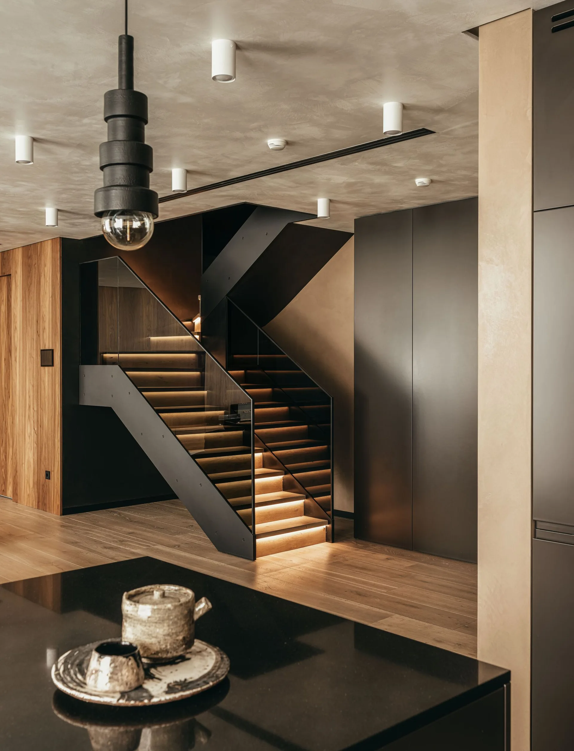 A modern wooden staircase with illuminated steps, a glass railing, dark matte cabinetry, and a textured plaster ceiling.