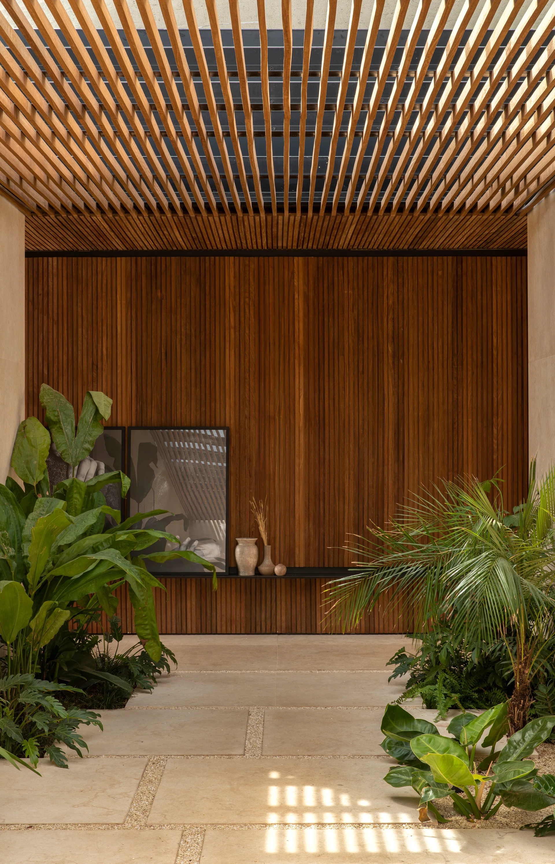 A modern courtyard features a slatted wood ceiling and matching wall cladding above stone pavers flanked by lush green plants.