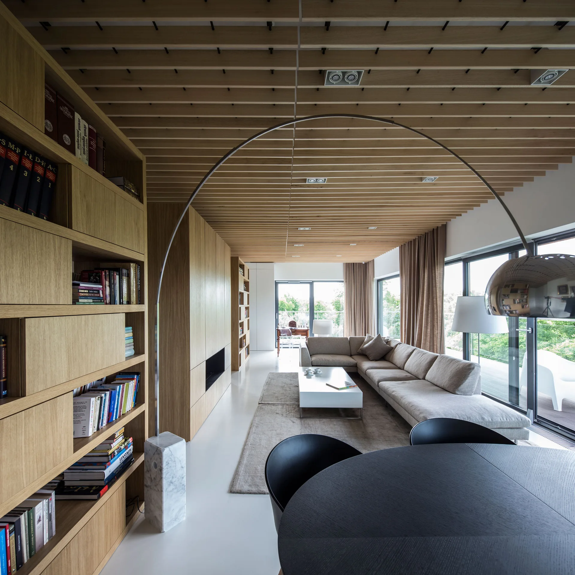 A modern living room featuring a light wood slatted ceiling, a large beige sectional sofa, a white coffee table, and built-in wooden bookshelves.