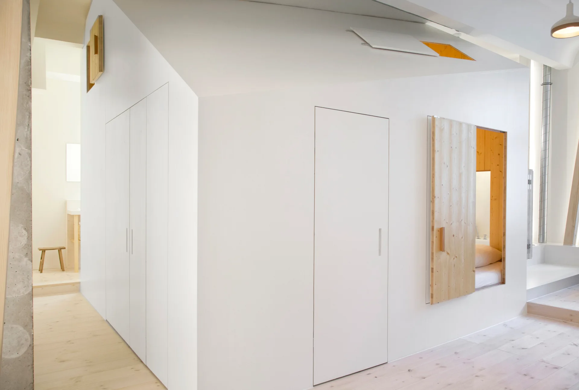 A minimalist white interior featuring flush-panel cabinetry, light wood floors, and a recessed wooden sleeping alcove with a sliding wood door.