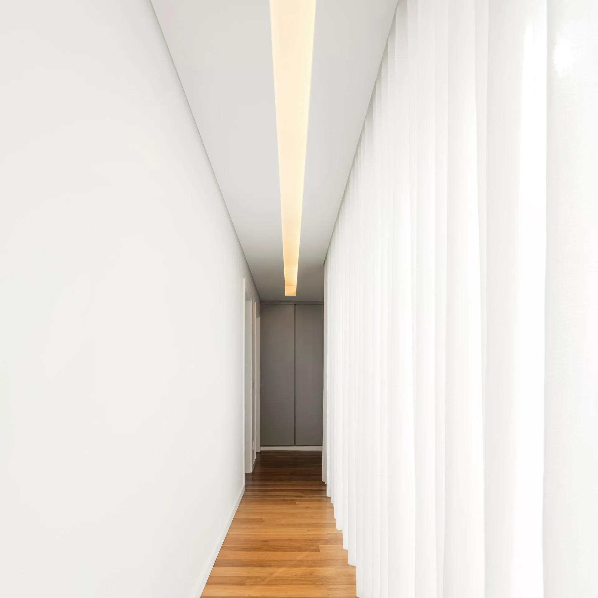 A minimalist hallway with wood plank flooring, plain white walls, and a wall of floor to ceiling white pleated curtains.