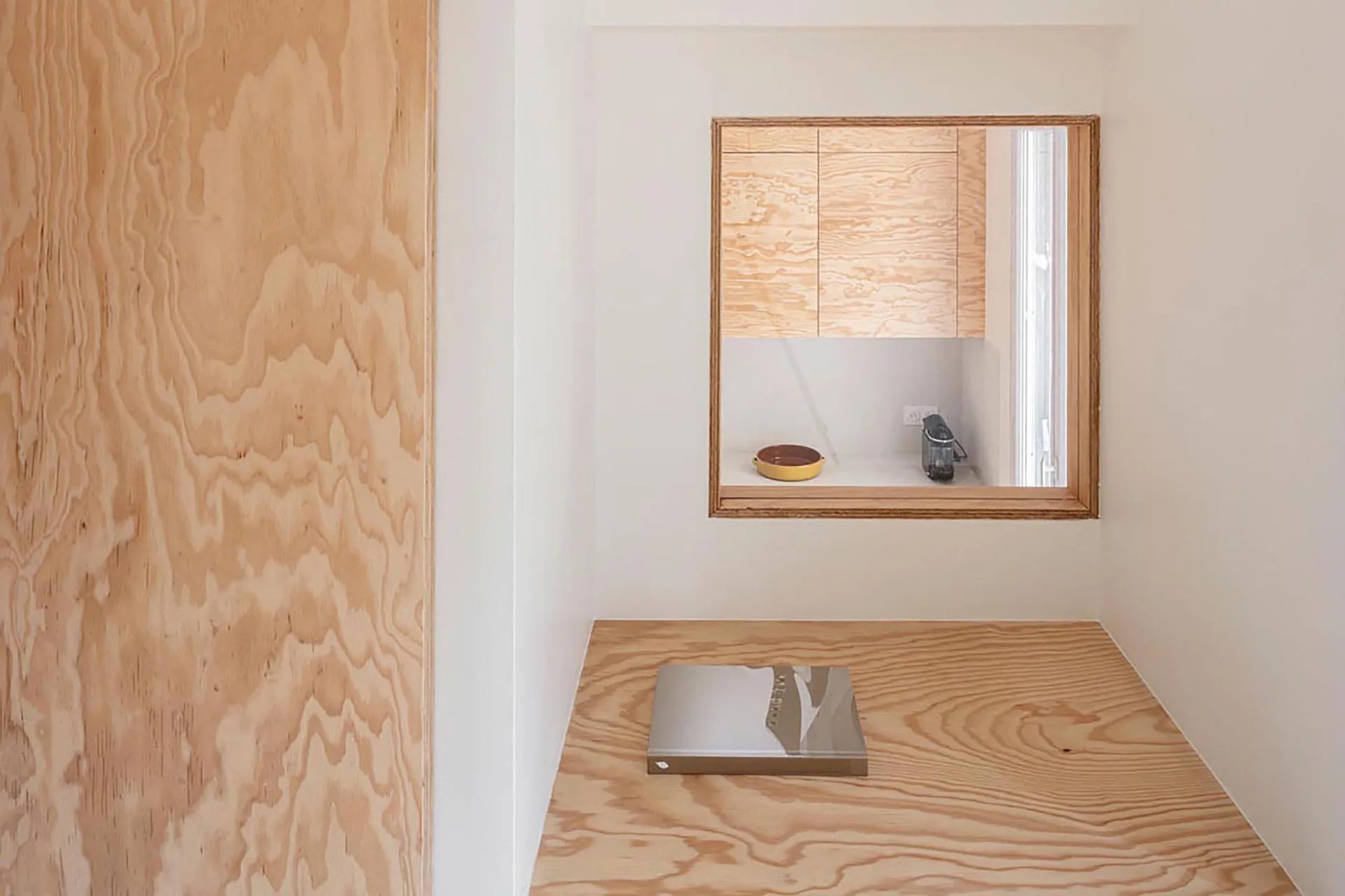 A minimalist white room features a raw plywood floor platform and a square interior window looking into a kitchen with matching plywood cabinetry.
