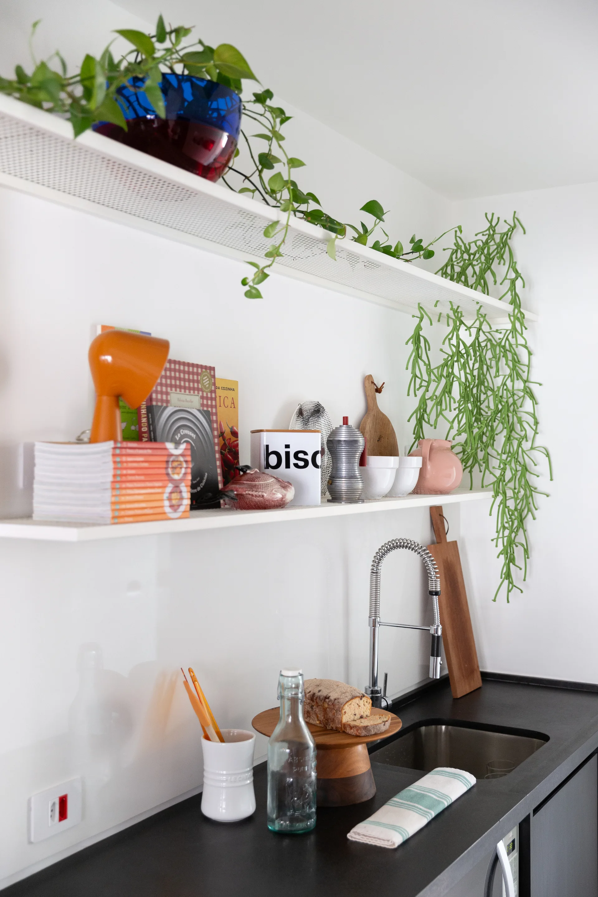 White perforated metal open shelves styled with plants, books, and kitchen accessories hang above a black countertop with an undermount sink.