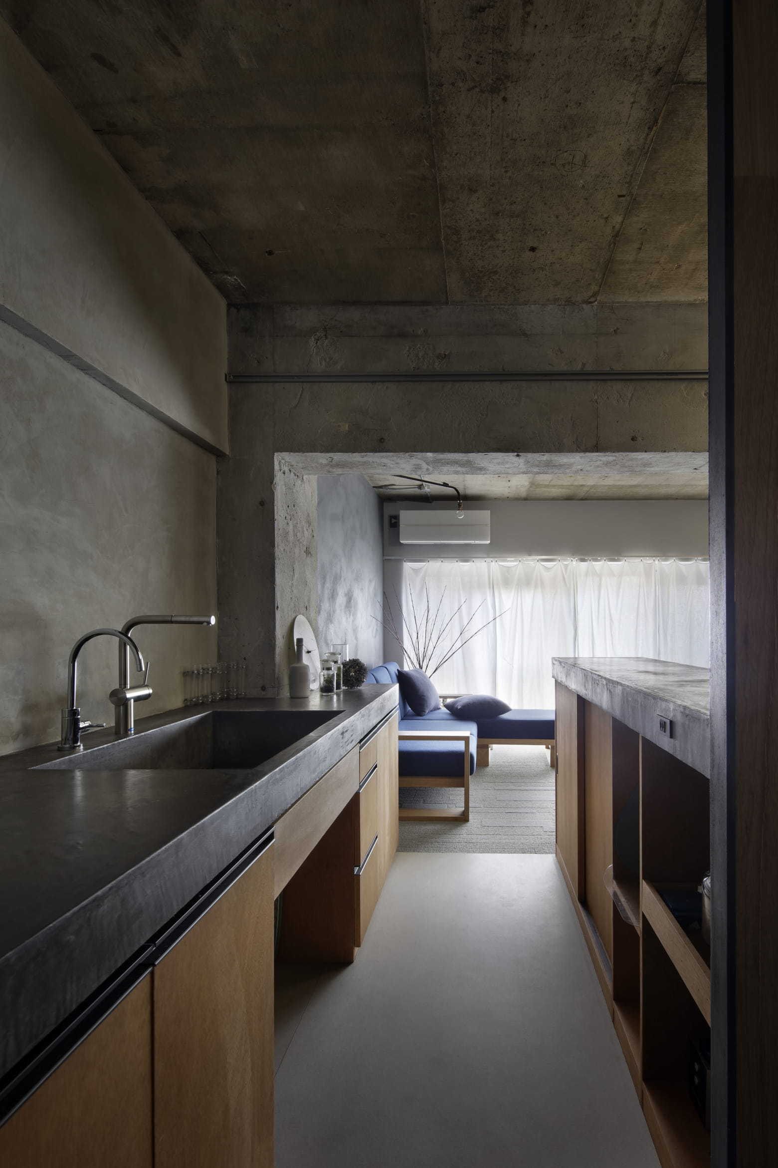 A galley kitchen featuring a thick concrete countertop and wooden cabinets leads into a living area with a blue sofa and sheer white curtains.