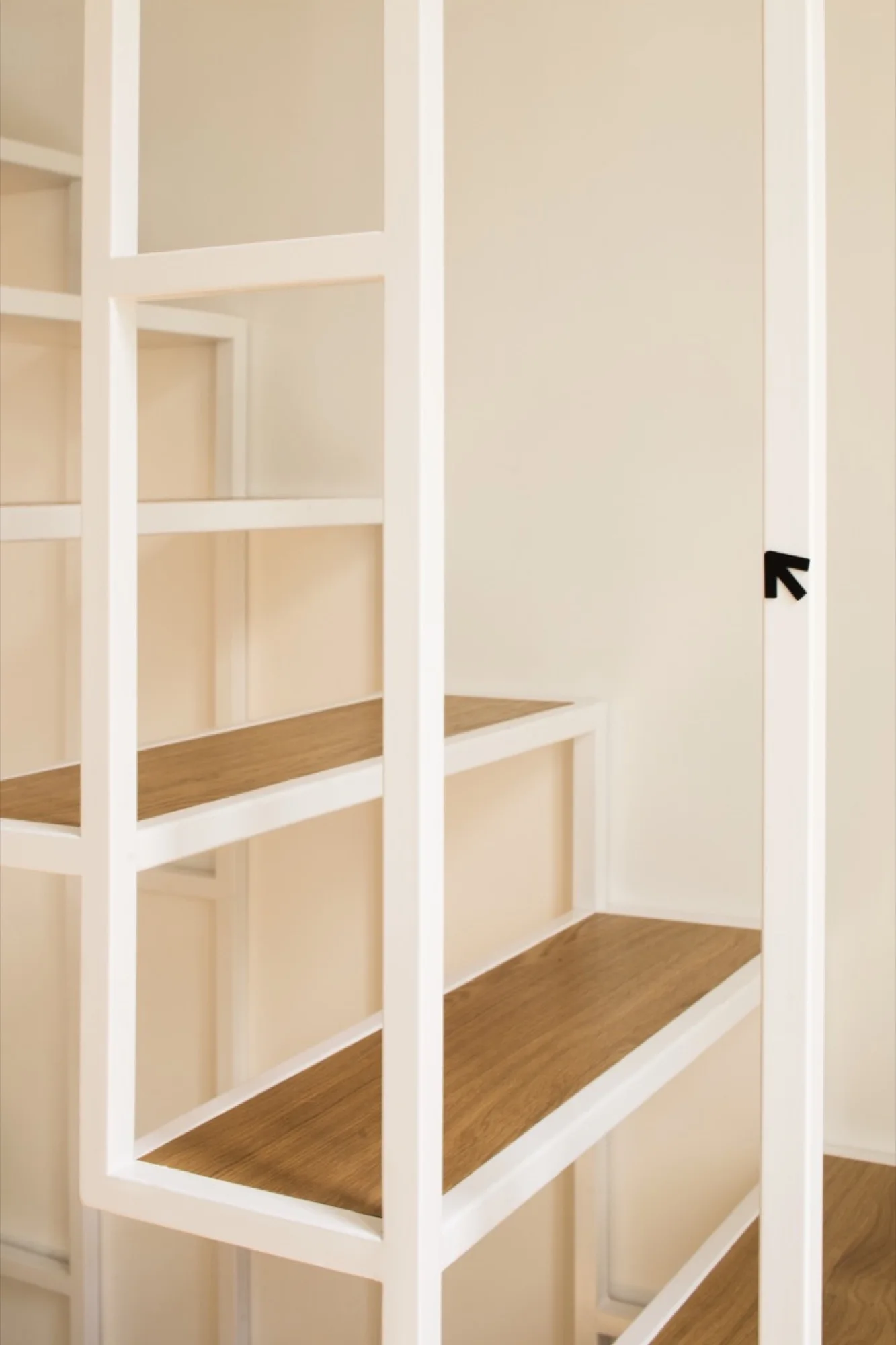 A modern staircase features warm wood treads supported by a geometric white metal frame set against an off-white wall.