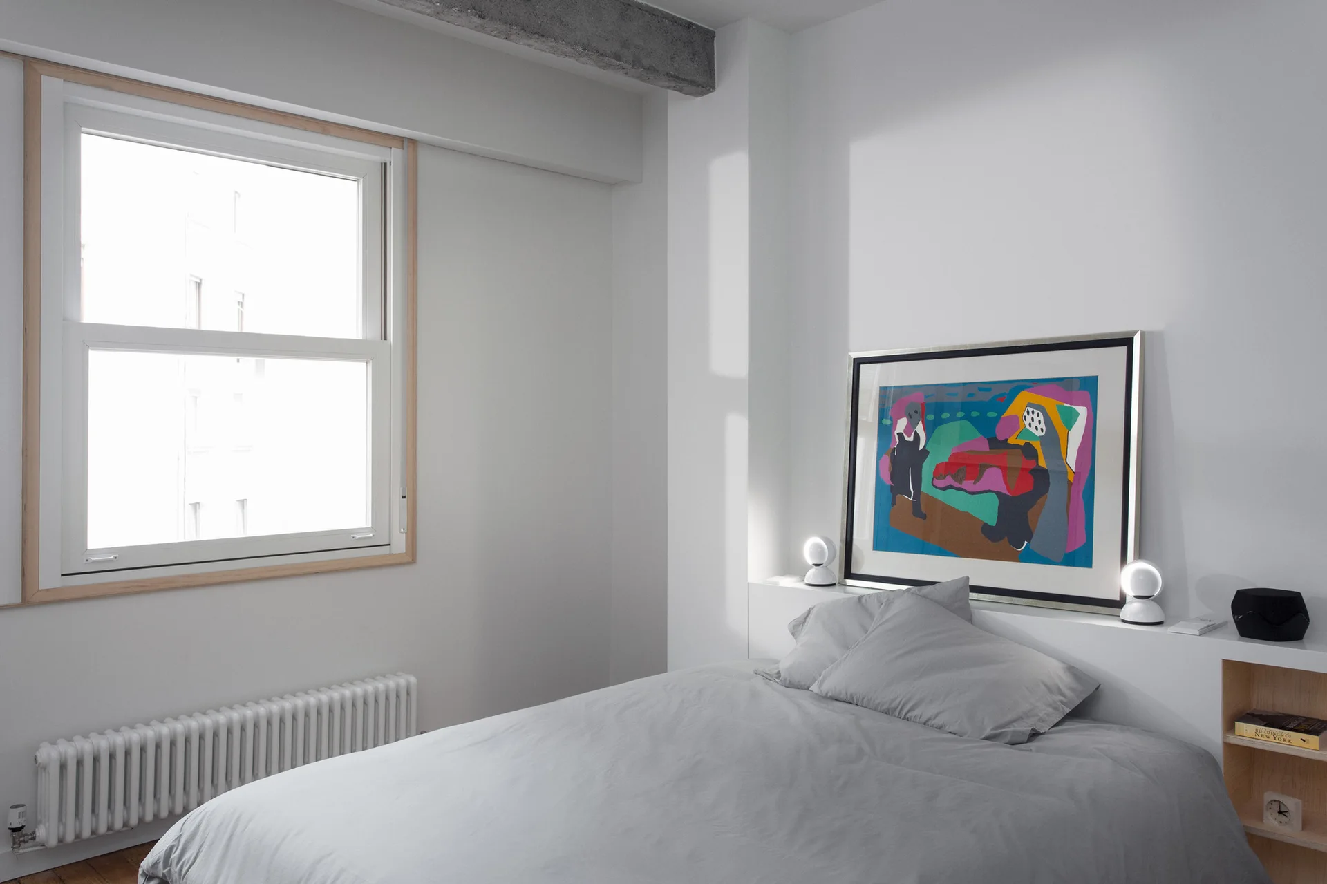 A minimalist bedroom features grey bedding, a low built-in white headboard ledge displaying framed art, and a wood-trimmed window.