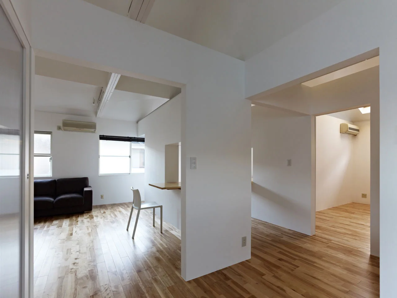 A minimalist living space features white walls, natural wood flooring, a dark sofa, and a small wall-mounted desk with a white chair.