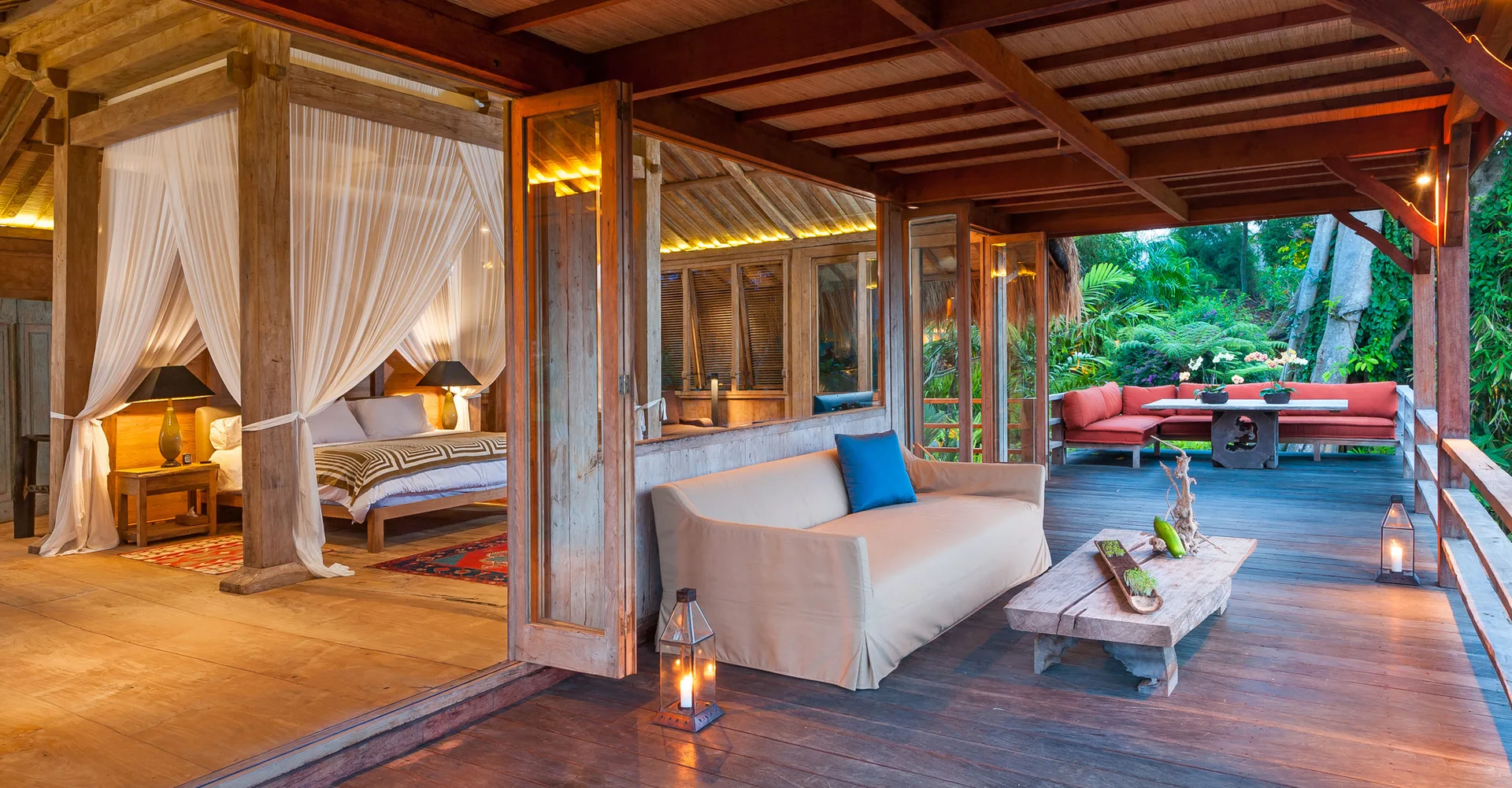 An open-concept tropical suite features a wood canopy bed with sheer white drapes next to a covered deck with a beige sofa.