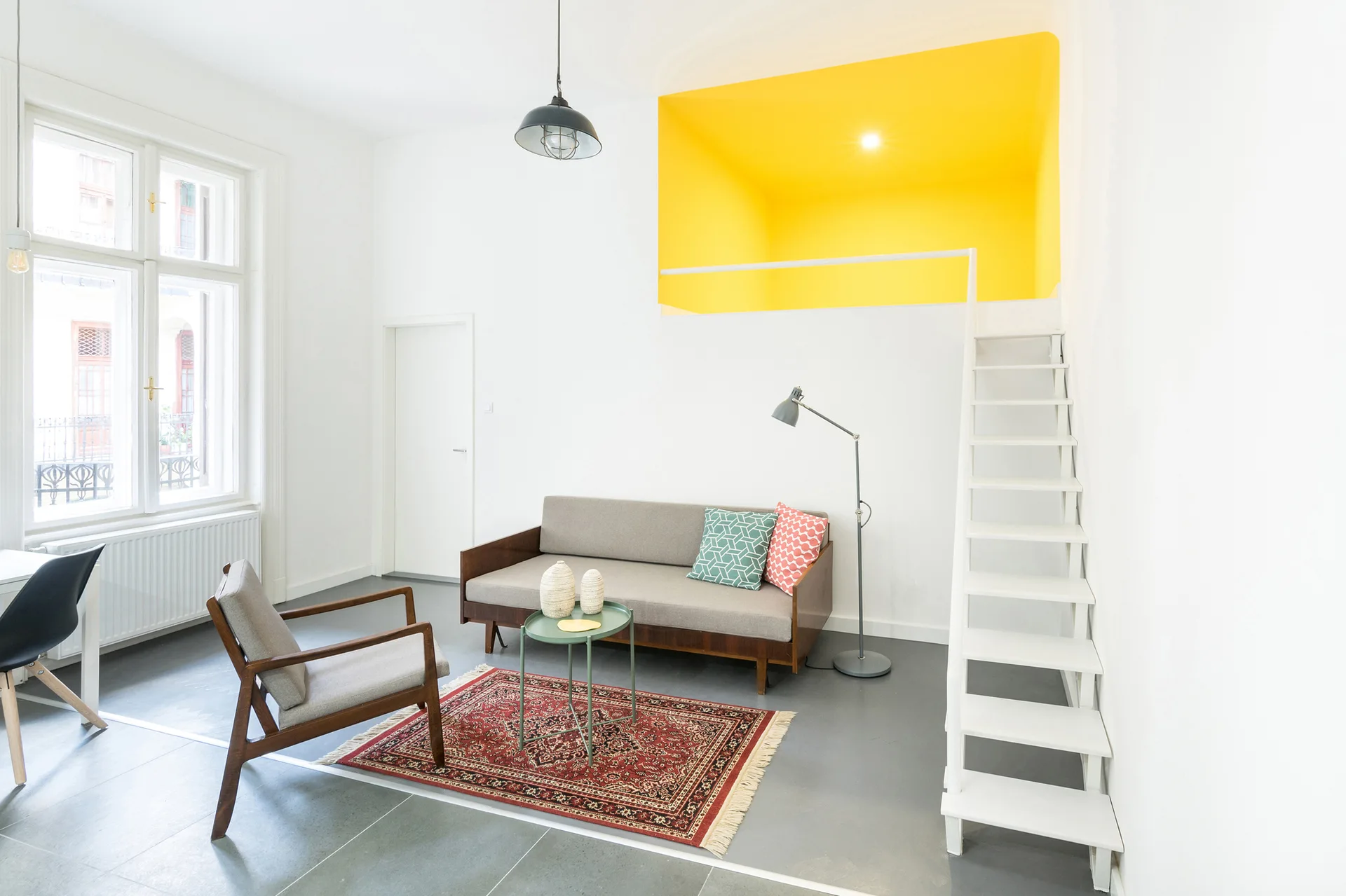 A modern white living room features a grey sofa, mid-century wooden chair, red patterned rug, and a white ladder leading to a bright yellow loft nook.