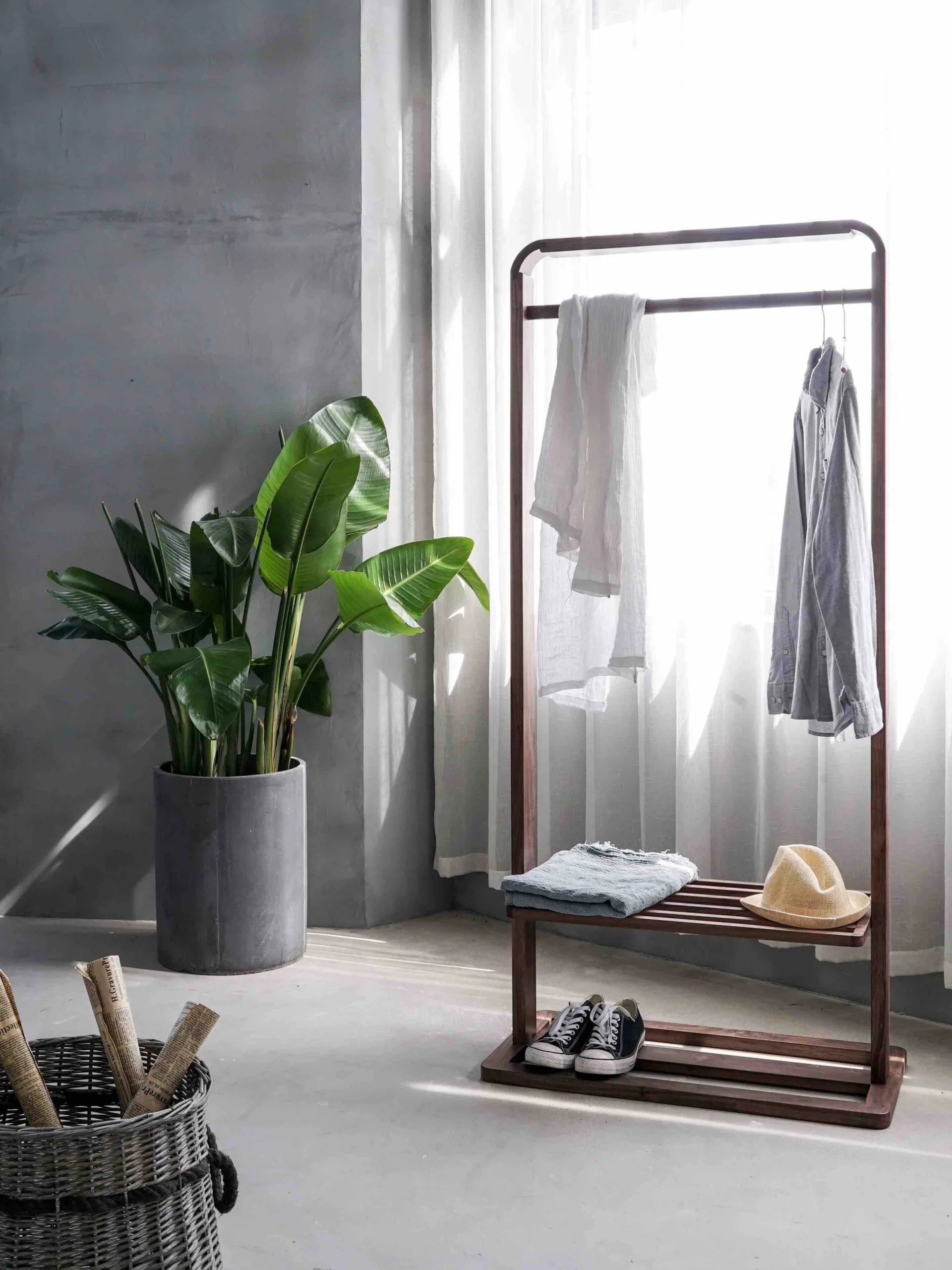 A wooden clothing rack holding a shirt and towel stands next to a large potted plant against a grey wall and sheer white curtains.