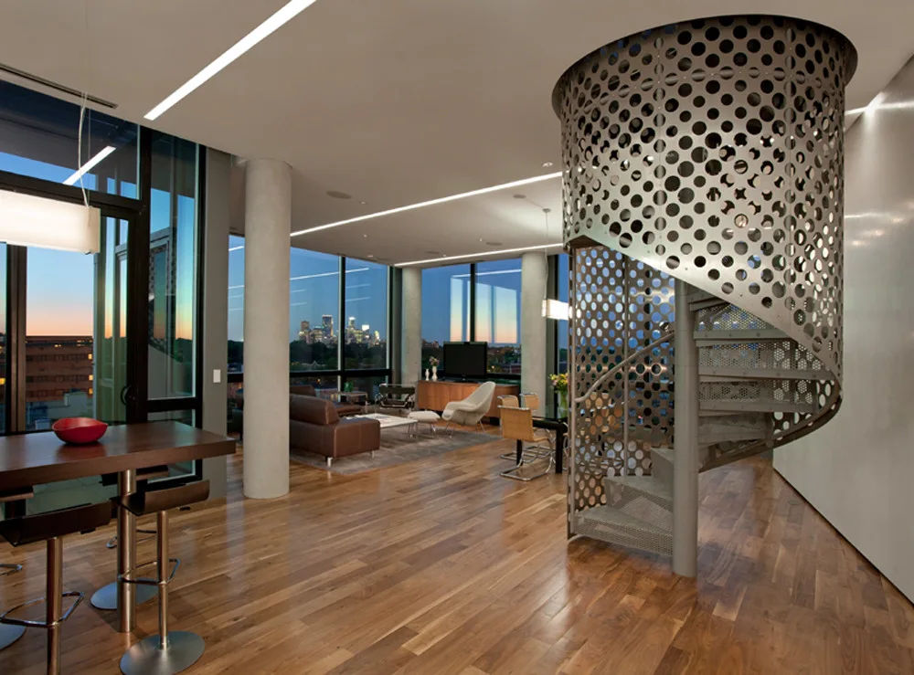 A modern open-plan living room features a perforated metal spiral staircase, hardwood flooring, and floor-to-ceiling windows showing a city skyline.